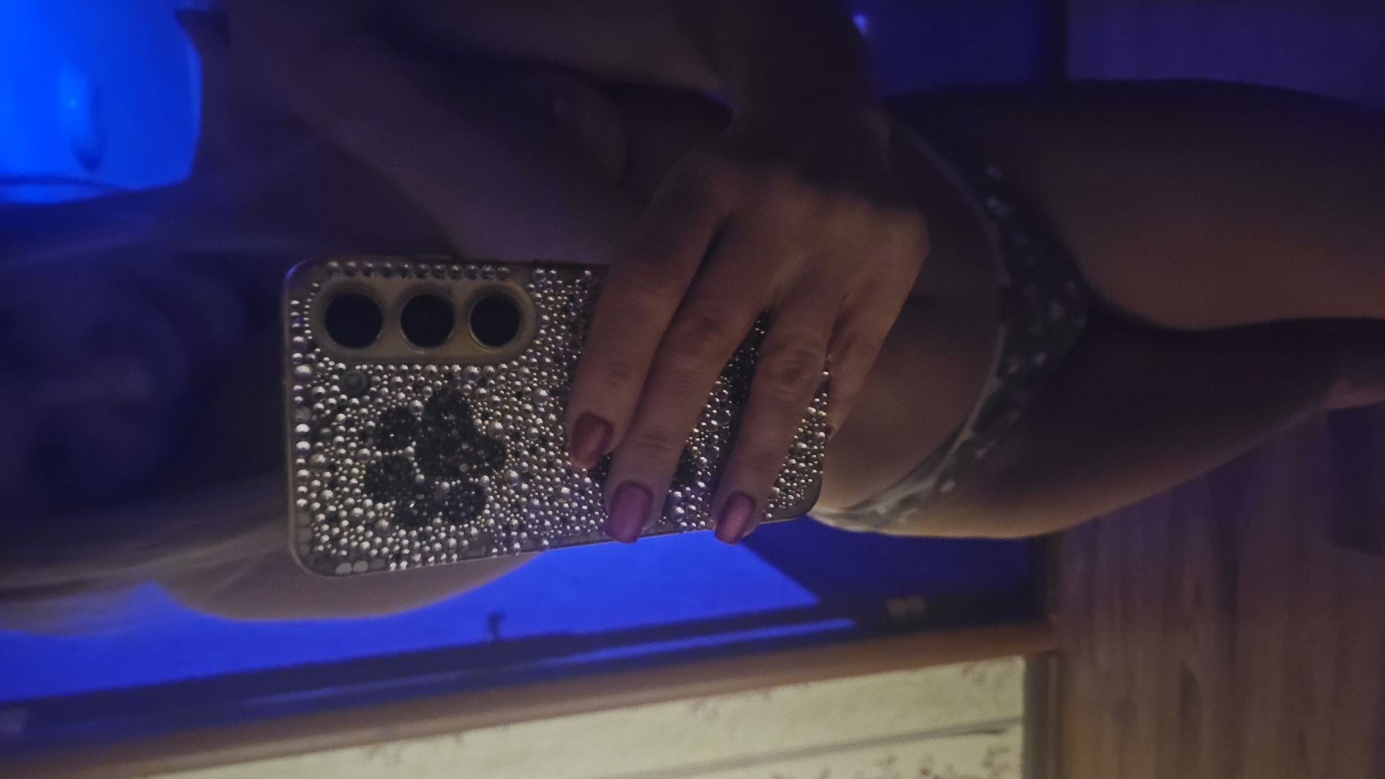 The image displays a mirror selfie taken indoors, rotated ninety degrees clockwise. A person's hand, holding a smartphone, is prominent in the center, with the fingers wrapped around the device. The fingernails are painted with a dark reddish-purple polish. The smartphone is encased in a highly decorative cover, embellished with numerous small, shiny, pearl-like studs, and features a distinct paw print design created from darker studs on its back. The phone's triple-lens camera array is visible through openings in the case.
Reflected in the mirror, parts of the person's body are visible, including their lower torso and upper legs, clad in patterned underwear, possibly a leopard or animal print. A strong blue light source emanates from the left side of the frame, casting a vibrant blue glow across the reflective surface and parts of the reflection. The overall lighting conditions are dim, with the blue light creating a distinct ambience. No specific location cues for Sevastopol, Ukraine, are visually present within the image, nor is any text visible. The activity depicted is the act of taking a photograph of one's own reflection in a mirror.