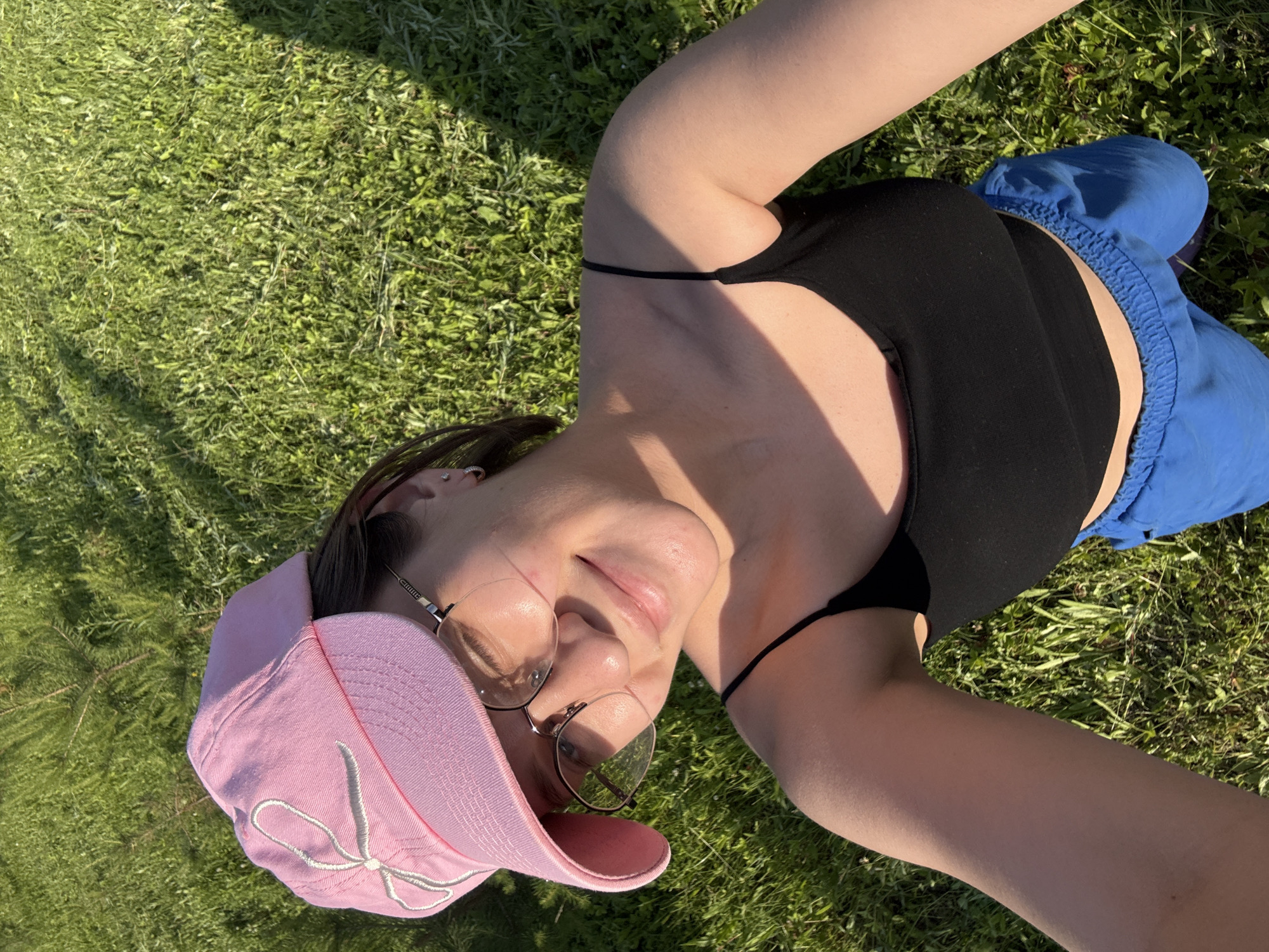 A young woman with dark hair, wearing a pink baseball cap with a white bow detail, glasses, a black tank top, and blue shorts, takes a selfie outdoors. She is smiling and looking at the camera. The background is a sunlit grassy area. There is no indication of the city of Doksha, Russia in this image.