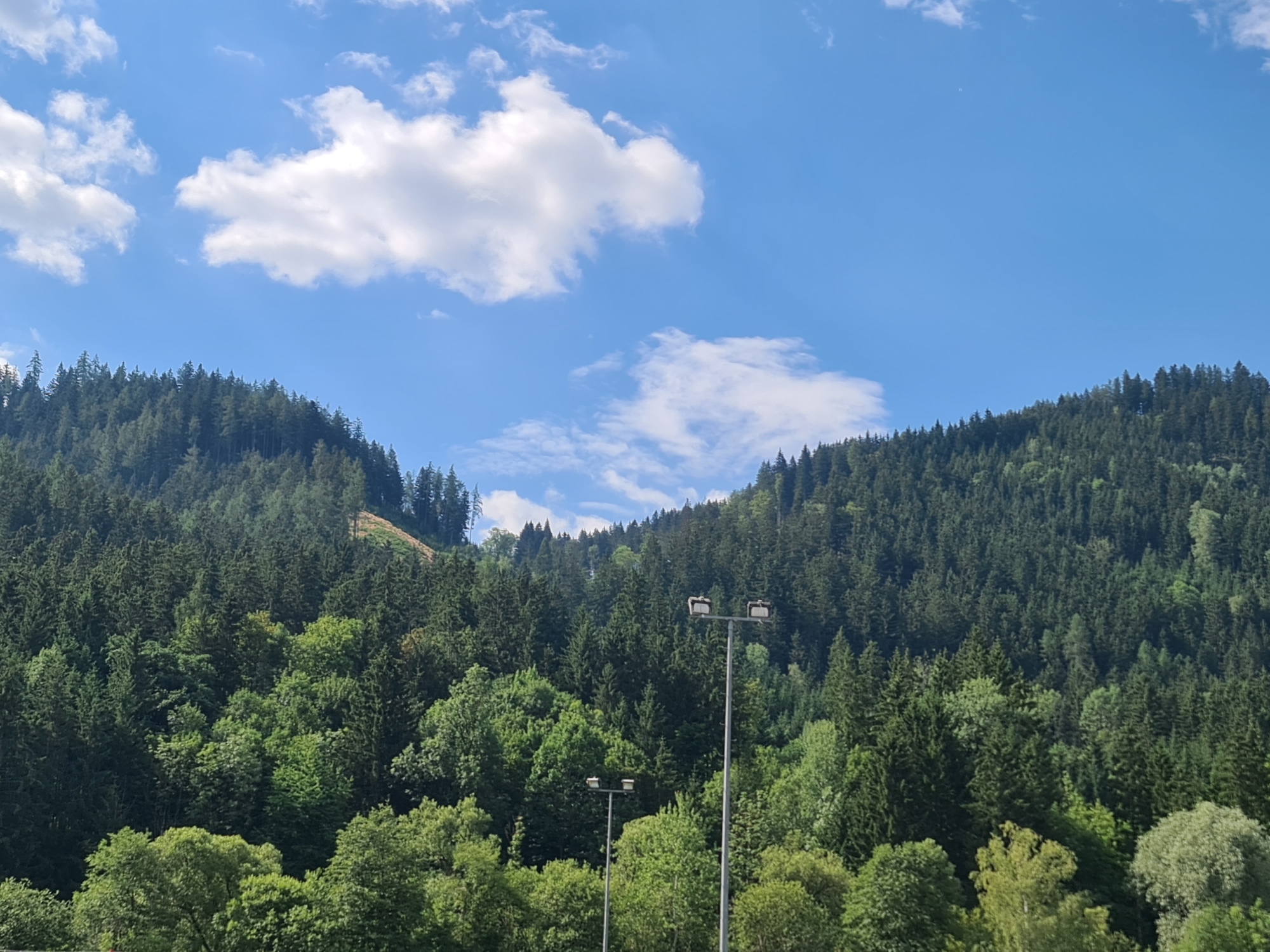 The image depicts a view of a forested hillside under a bright blue sky with scattered white clouds. Two tall lamp posts with multiple lights are visible in the foreground, positioned in front of the dense evergreen forest that covers the slopes. The trees are lush and varied in shades of green. In the distance, the hillside shows a patch cleared of trees, revealing lighter soil. The scene is serene and natural, suggesting a location within or near a natural park or recreational area. This scene is located in Sankt Marein im Mürztal, Austria.