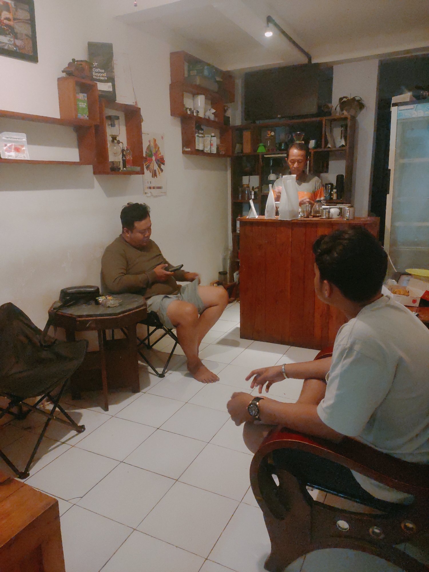 In the city of Sooko, Indonesia, a coffee shop scene unfolds. Two men are present. One man, seated on a folding chair, is holding a mobile phone and looking down at it. He is wearing a brown long-sleeved shirt and grey shorts. Adjacent to him is a hexagonal table with a dark-colored bag and what appears to be a pack of cigarettes. Another man stands behind a wooden counter, possibly a barista, interacting with something on the counter. He is wearing an orange and grey striped shirt. The background reveals shelves filled with various items, including coffee-making equipment, jars, and decorative objects. The floor is tiled with white square tiles. The lighting suggests an indoor setting, likely during the evening.