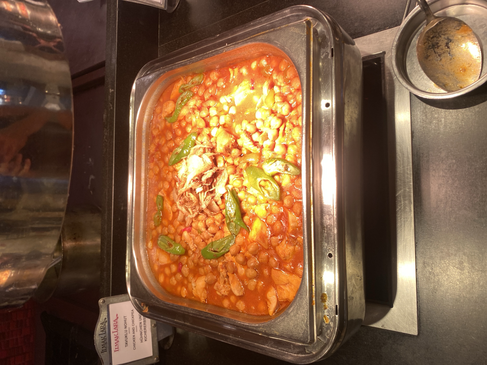 A chafing dish filled with a hearty chickpea and chicken stew is presented in a buffet setting. The stew is a rich, reddish-brown color, with visible chickpeas, chunks of chicken, and sliced green peppers. The dish is garnished with a small pile of crispy fried onions on top. The chafing dish itself is made of stainless steel and rests on a metallic base. To the left of the chafing dish, a small sign with the logo "ILMAK LARA" and text in Turkish and English reads "Tavuklu Nohut Chicken and Chickpea" and "Hühnchen und Kichererbs". A serving spoon, also showing signs of use, rests in a small metal holder to the right of the chafing dish. The background suggests a commercial establishment with polished metal surfaces and dark countertops. The lighting is warm, likely from overhead buffet lights, creating reflections on the metallic surfaces. The image is taken from a slightly elevated, forward-facing perspective. The specific location in Aksu, Turkey, is not discernible from the visual information provided.