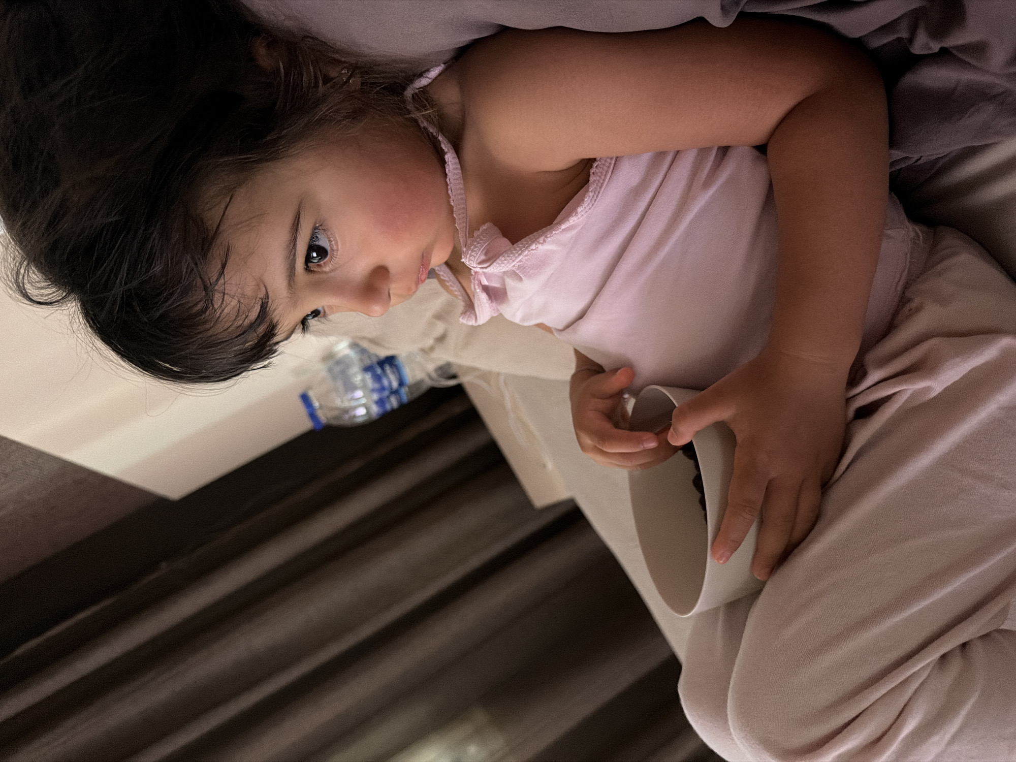 A young girl with dark hair is lying in bed, holding a white mug. She is wearing a light pink tank top and light-colored pants. Her face is turned to the side, and she has a thoughtful expression. A partially visible bottle of water is on a surface next to her. The background shows a bed with gray and light pink bedding, and dark, ribbed curtains. The lighting suggests it might be nighttime or an indoor setting with low light. The image appears to have been taken in Burgas, Bulgaria.