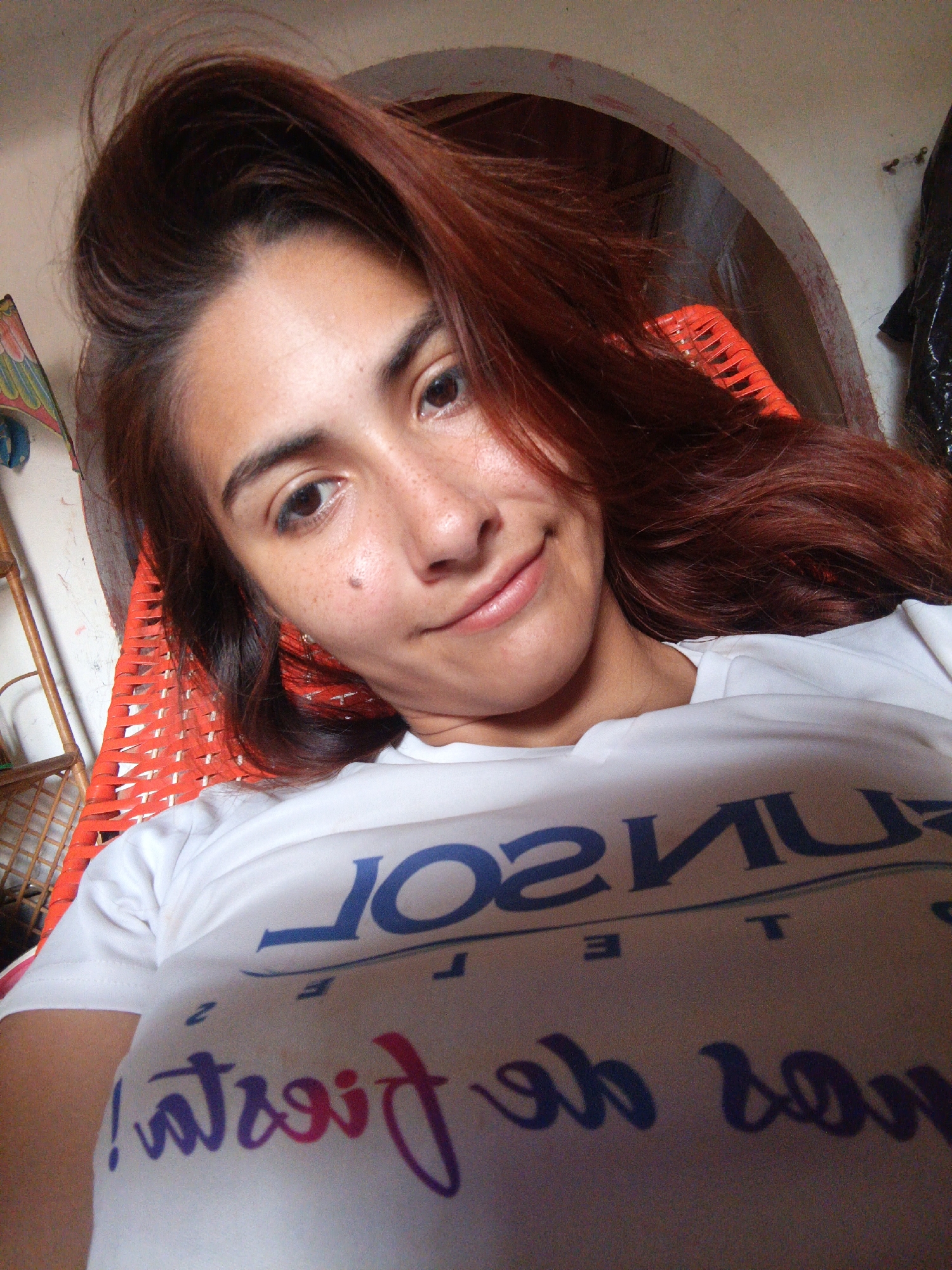 A young woman with auburn hair and freckles is taking a selfie in a casual, outdoor setting. She is lying down, possibly in a hammock or reclining chair, as indicated by the orange woven material visible behind her. The environment appears to be outdoors, with hints of a rustic structure, possibly an arched doorway, in the background. The woman is wearing a white t-shirt with a logo and text printed on it. The text on the shirt is partially obscured and appears to be in blue and purple lettering. There are no other people or animals visible in the image. The lighting suggests it is daytime. The location is identified as Juan Griego, Venezuela.