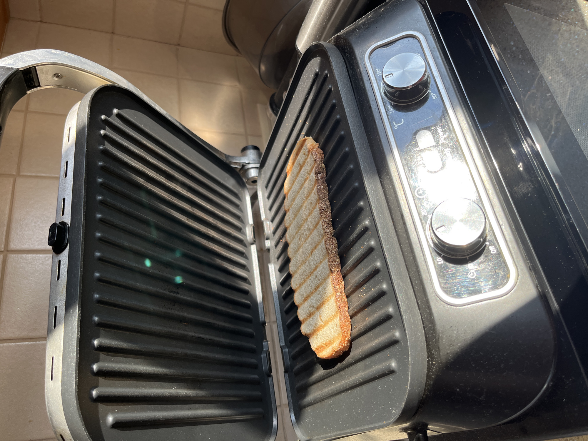 A close-up, eye-level shot shows a slice of bread being toasted on an open panini press. The bread is golden brown with characteristic grill marks and is positioned horizontally on the bottom plate of the press, which has a non-stick, ribbed surface. The panini press is black with silver accents and features two control dials and a light indicator. The upper plate of the panini press is open, revealing the ribbed interior. The background consists of a tiled wall, suggesting the appliance is in a kitchen environment. The lighting indicates the presence of natural sunlight, casting shadows on the appliance. There are no people visible in the image. The scene is devoid of any indication of the city of Balashikha, Russia, or any specific geographic location beyond a generic kitchen setting.