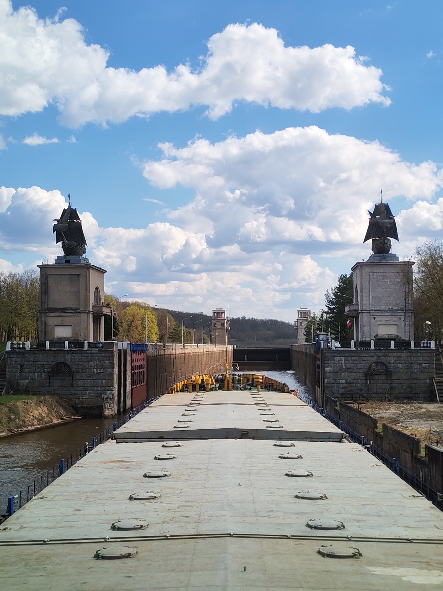 The image shows a wide, flat-topped barge positioned in a lock on the Moscow Canal, in Yakhroma, Russia. The barge fills the width of the lock. Above the lock are two towers, each topped with a sculpture of a sailing ship. In the background are two more towers, possibly control towers for the lock. The sky is blue with scattered white clouds. Green trees line the canal banks on either side of the lock.