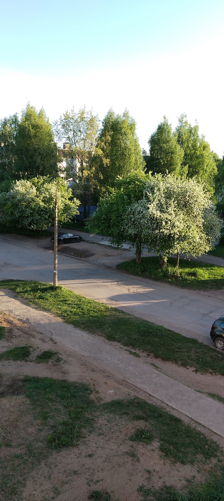The image depicts a street scene in Chaikovsky, Russia, captured from an elevated perspective. The scene includes a network of roads and pathways, interspersed with patches of grass and trees. A light pole stands near the intersection of the roads. Two cars are visible: a dark-colored sedan parked along the road and another dark-colored vehicle parked further down the road. Several buildings are visible in the distance, partially obscured by trees. The sky is clear and light blue, suggesting a sunny day. The trees are green and lush, with some showing signs of flowering, indicating it may be spring.