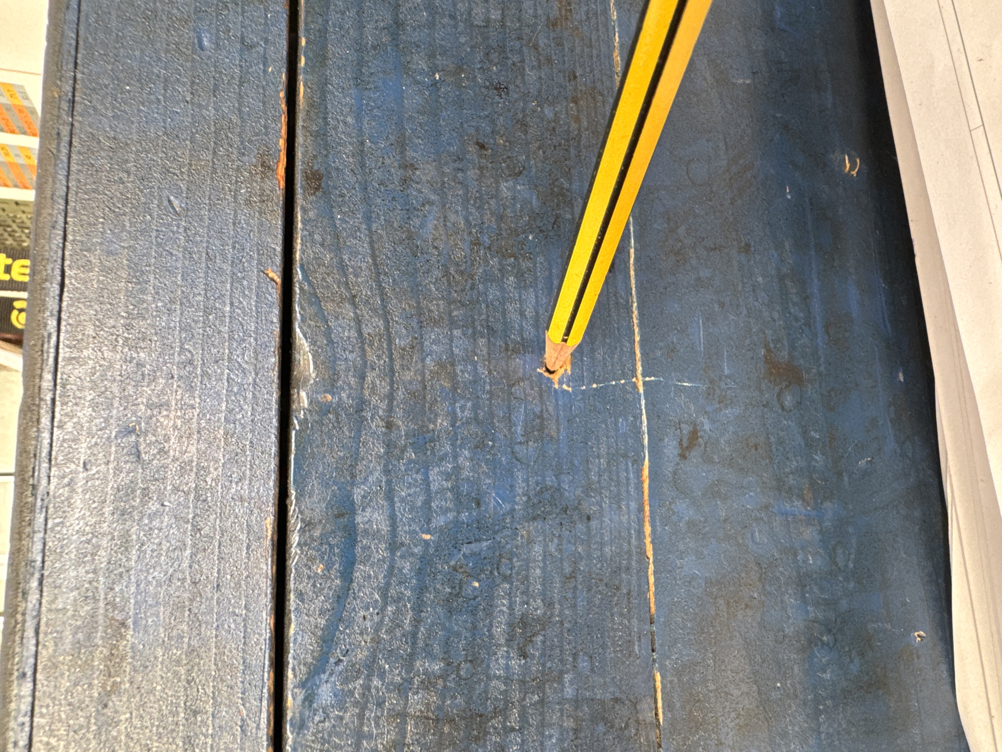 A close-up, overhead shot of a weathered, blue wooden surface shows a yellow pencil with a black stripe angled across the lower right portion of the frame. The pencil tip is touching a small, shallow indentation in the wood, suggesting it was used to mark or poke the surface. The wood appears to be constructed from planks, with visible grain lines and signs of wear, including chipped paint and minor discoloration. The background of the image is out of focus, but suggests some printed material or the edge of another object at the top and a white surface at the bottom. There are no people or any other identifiable objects present in the immediate scene. The image does not contain any information related to the city of Mejaniga, Italy.
