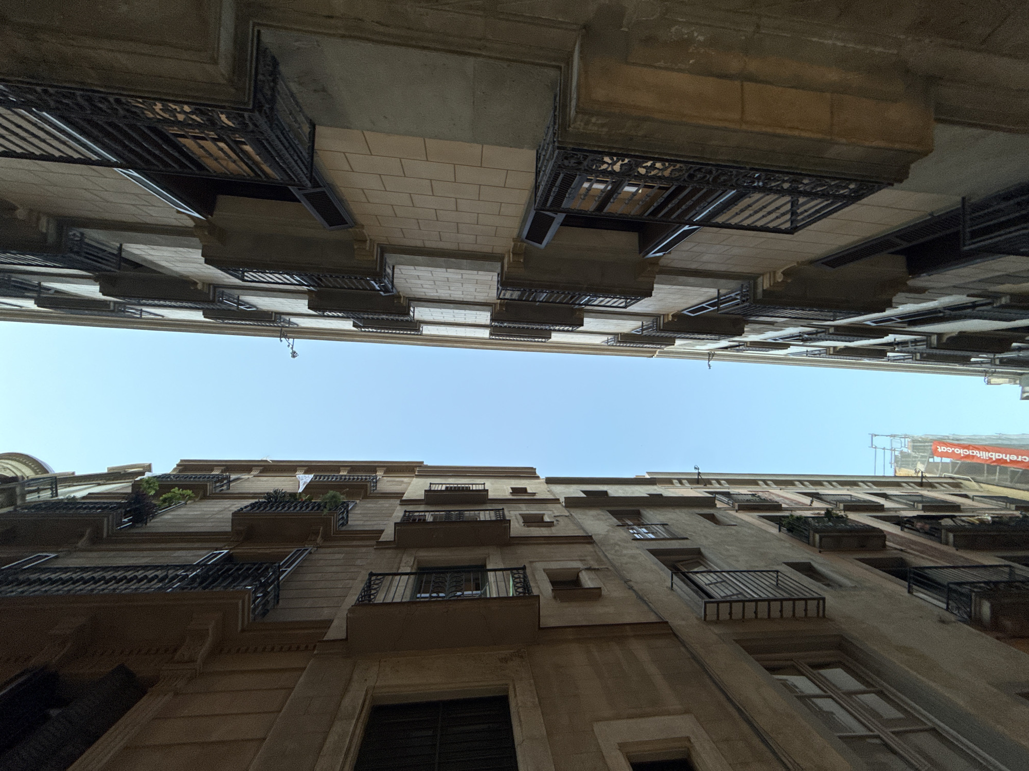 The photograph depicts a narrow street in Barcelona, Spain, shot from a low angle looking upwards. Buildings line both sides of the street, their facades featuring multiple stories with balconies. The structures are constructed of stone or concrete, exhibiting a weathered appearance. Some balconies are adorned with plants, while others display decorative ironwork. A construction banner, visible on one of the buildings, indicates ongoing renovation or restoration work. The sky is visible as a narrow strip between the buildings, suggesting a clear day. The composition emphasizes the height and density of the urban environment.