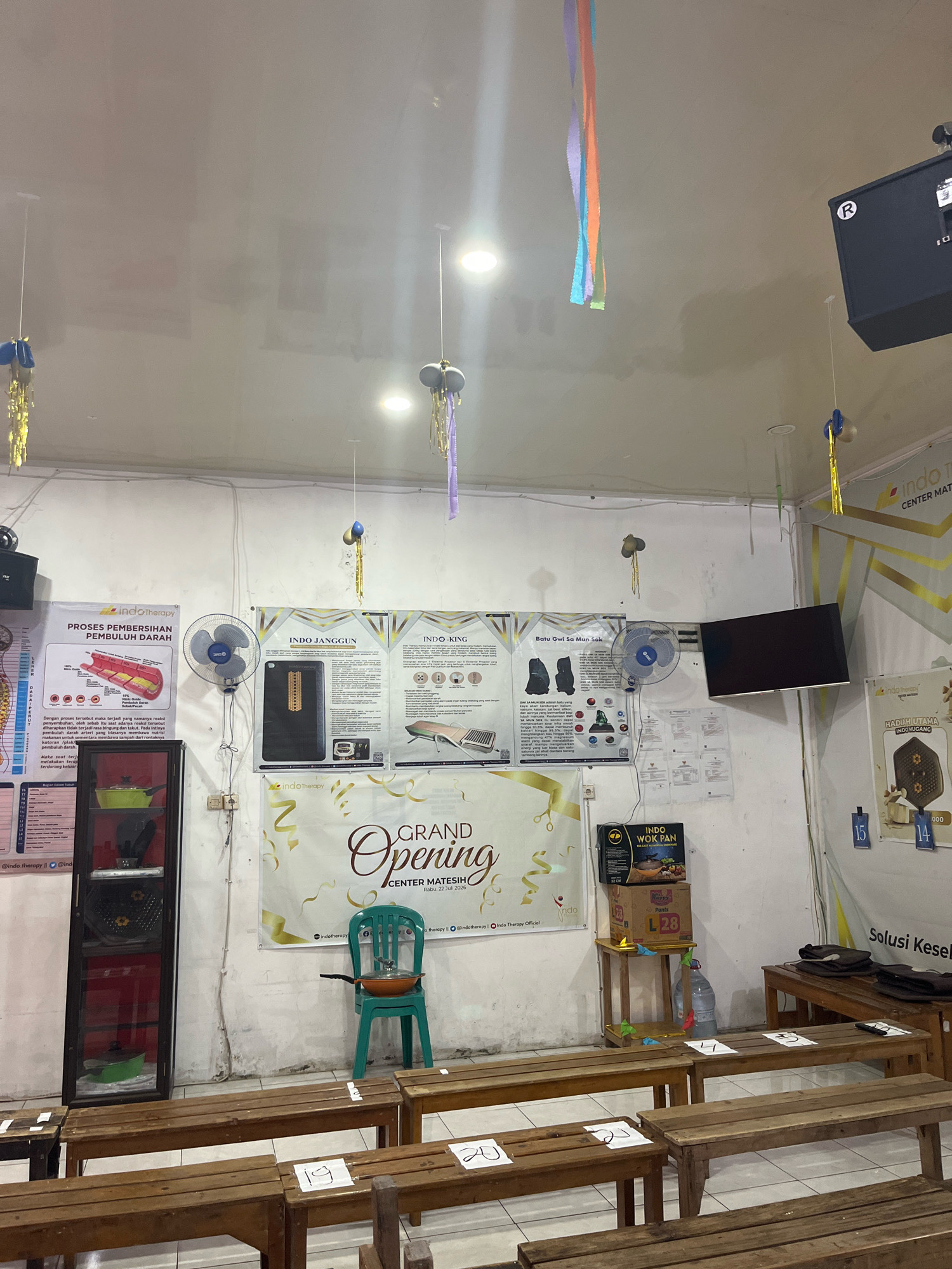The image depicts an indoor scene, likely a waiting area or event space, in Matesih, Indonesia. The room is sparsely furnished with rows of wooden benches. A large banner, visible on the back wall, announces "GRAND OPENING CENTER MATESH" with the date "Rabu, 22 Juli 2026". Various posters with text and diagrams are mounted on the walls, some relating to "Indo Therapy" and others with product information. A green plastic chair with a frying pan on its seat is positioned near the banner. Decorative streamers hang from the ceiling. Several fans are mounted on the walls, and a television screen is mounted higher up. A large black speaker is visible on the upper right. A tall glass-fronted cabinet containing various items, including a green pot, stands on the left side of the room. Boxes and a water bottle are placed on a wooden stand. Numbers on white paper are taped to the benches, suggesting some form of seating arrangement or queuing system. The overall impression is of a functional, perhaps community-oriented space prepared for an event.