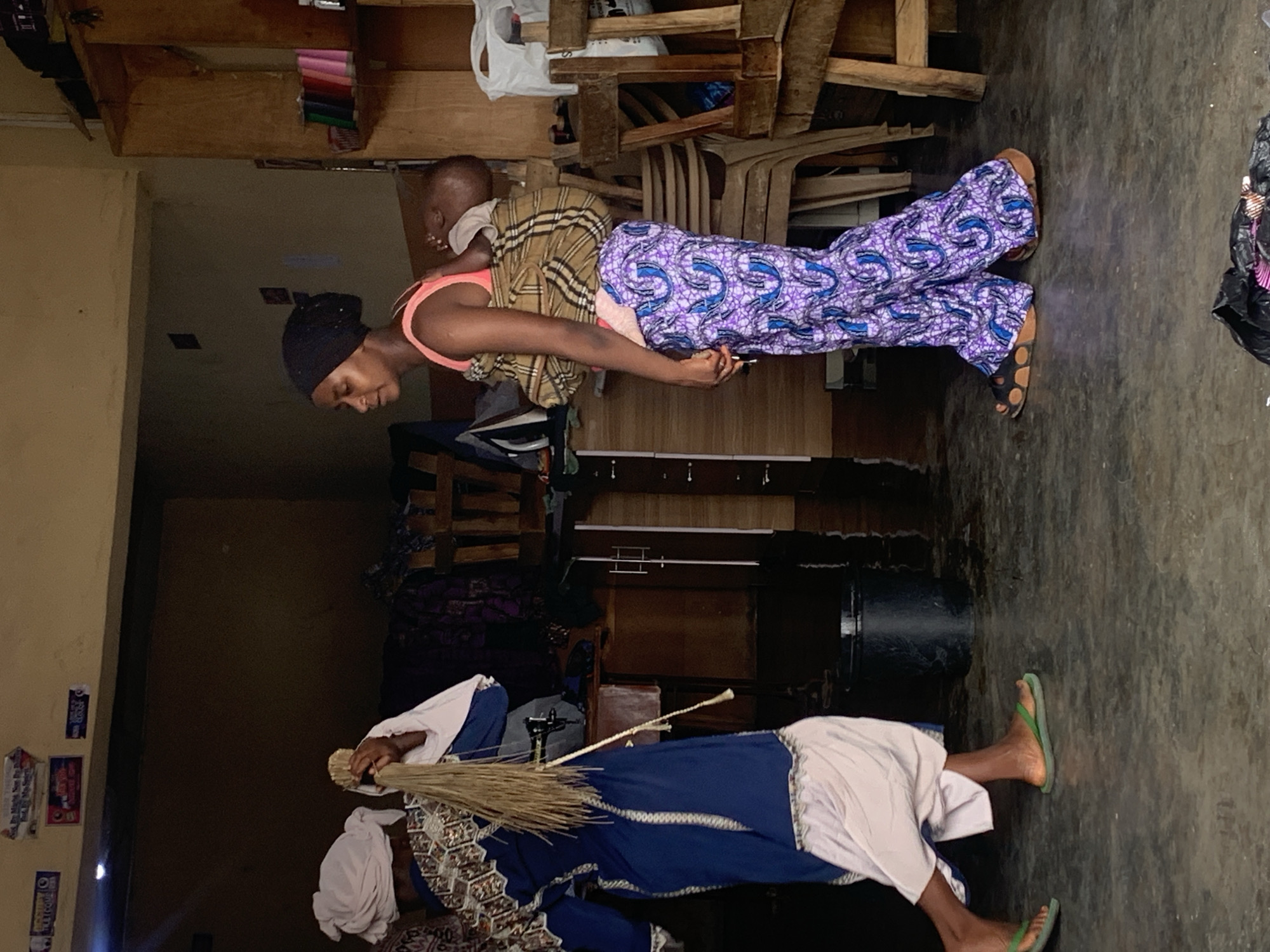 In this media file, two individuals are depicted within what appears to be an indoor setting, possibly a market or storage area, given the presence of shelves laden with various items and wooden structures. The scene is dimly lit, suggesting it might be evening or an interior with limited natural light.
The primary subject is a woman carrying a baby on her back. She is wearing a black head covering and an orange top, with a vibrantly patterned purple and blue fabric wrapped around her legs. The baby, strapped to her back with a patterned cloth, is looking away from the camera. The woman's expression is focused downwards.
In the foreground, another individual, dressed in a blue garment and a white head covering, is partially visible. This person is holding a broom made of straw. Their stance suggests they might be preparing to sweep or are in the process of cleaning. They are wearing green flip-flops.
The overall environment includes concrete flooring and wooden shelves filled with what seem to be textiles, rolls of material, and other stored goods. The presence of a large black bucket is also notable.
There is some visible text on the wall in the background, appearing as small posters or labels, but it is too small and blurry to be legible. The image has a candid feel, capturing everyday life in what is indicated to be Jos, Nigeria. The lighting and the activity suggest a moment captured during a potentially busy or routine part of the day.