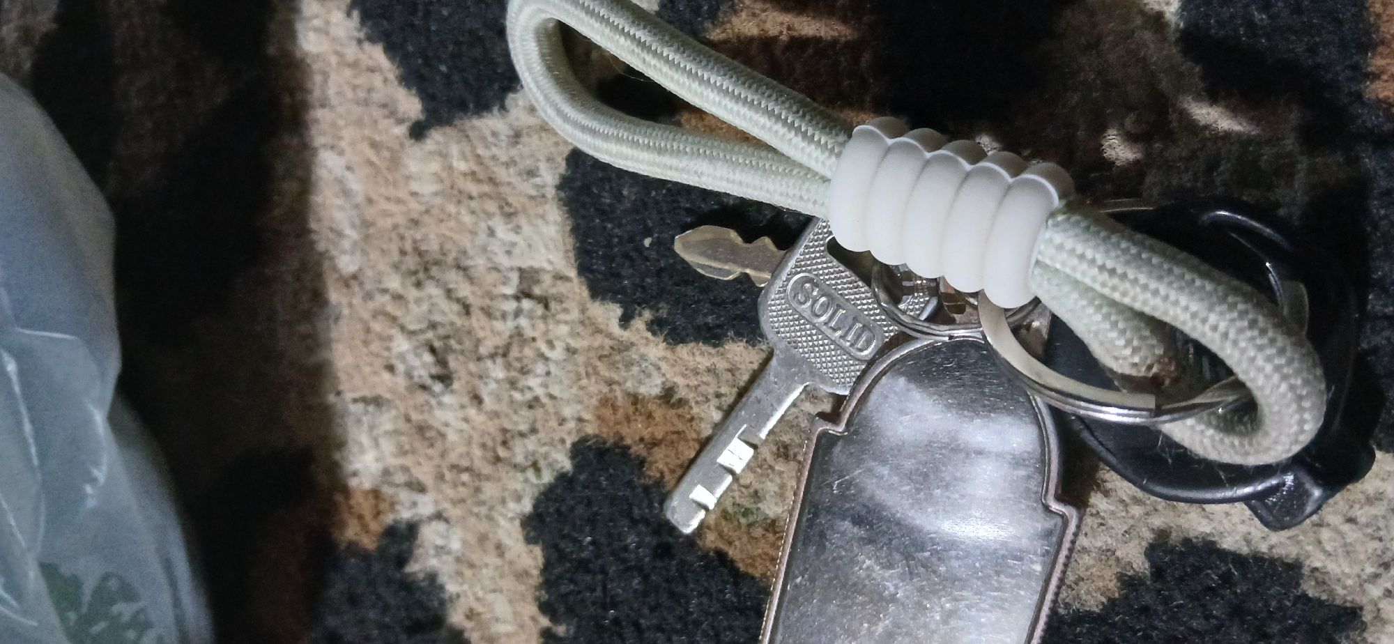 In Demangan, Indonesia, a close-up shot focuses on a keychain with several items attached. The keychain features two metallic keys, one of which is partially obscured and has the word "SOLID" embossed on it. There is also a large, metallic tag or plate, possibly a luggage tag, with a smooth, reflective surface. The keys and tag are connected to a light-colored, braided cord that is secured by a white, ribbed plastic fastener. The background consists of a textured surface with a camouflage-like pattern of black, brown, and beige. The lighting is dim, casting shadows and highlighting the metallic sheen of the keys and tag.