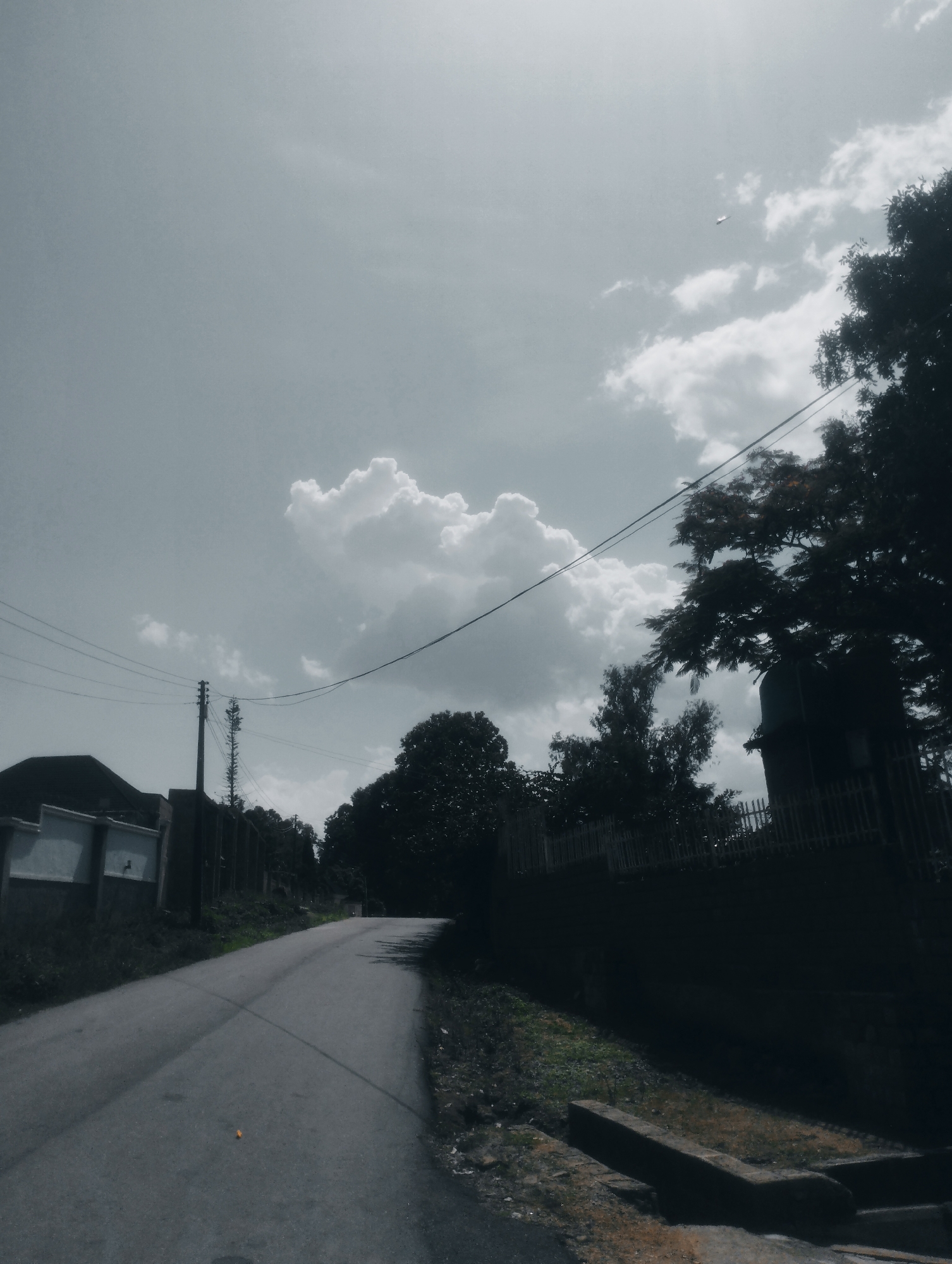 A drone flies in the sky above a road in Jos, Nigeria. The sky is filled with clouds, and the sun is shining. The road is asphalt and curves to the left. There are trees and buildings on either side of the road. The trees are green and lush, and the buildings are made of concrete. The drone is a small, white object that is visible in the sky. The drone is flying at a high altitude, and it is difficult to see the details of the drone. The drone is a commercial drone, and it is being used for aerial photography. The drone is a DJI Mavic Pro.