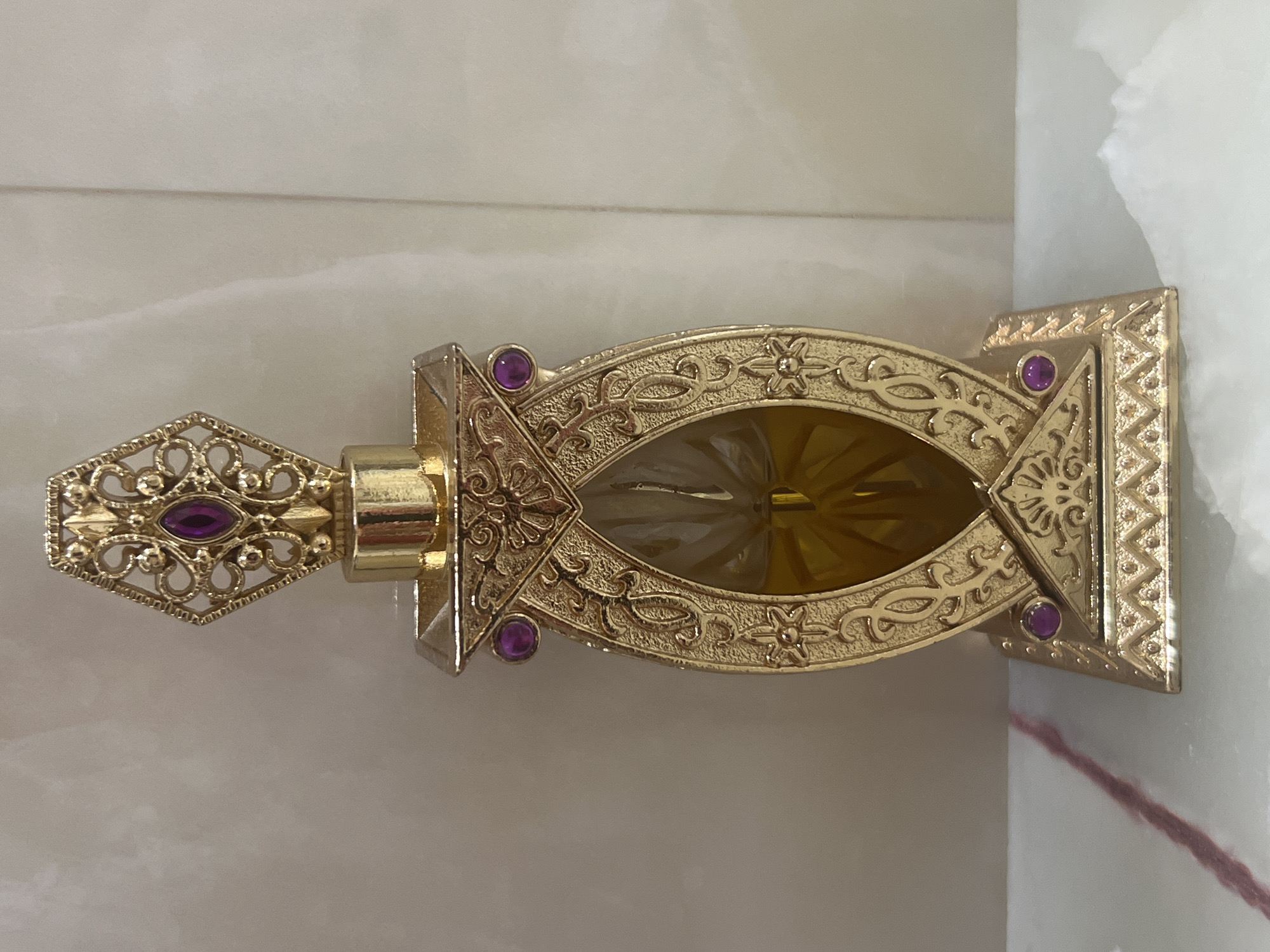 A gold-colored perfume bottle with intricate metalwork and purple gems is displayed on a marble surface. The bottle is elongated and oval-shaped, with a decorative cap featuring a large purple gem at its apex. The body of the bottle is adorned with ornate gold filigree and smaller purple gems on either side. The liquid inside the bottle appears to be yellow. The background is a textured, light-colored wall, possibly marble or a similar stone. The lighting is even, highlighting the details of the bottle. There are no people present in the image, nor any indication of the city of Bender, Moldova.