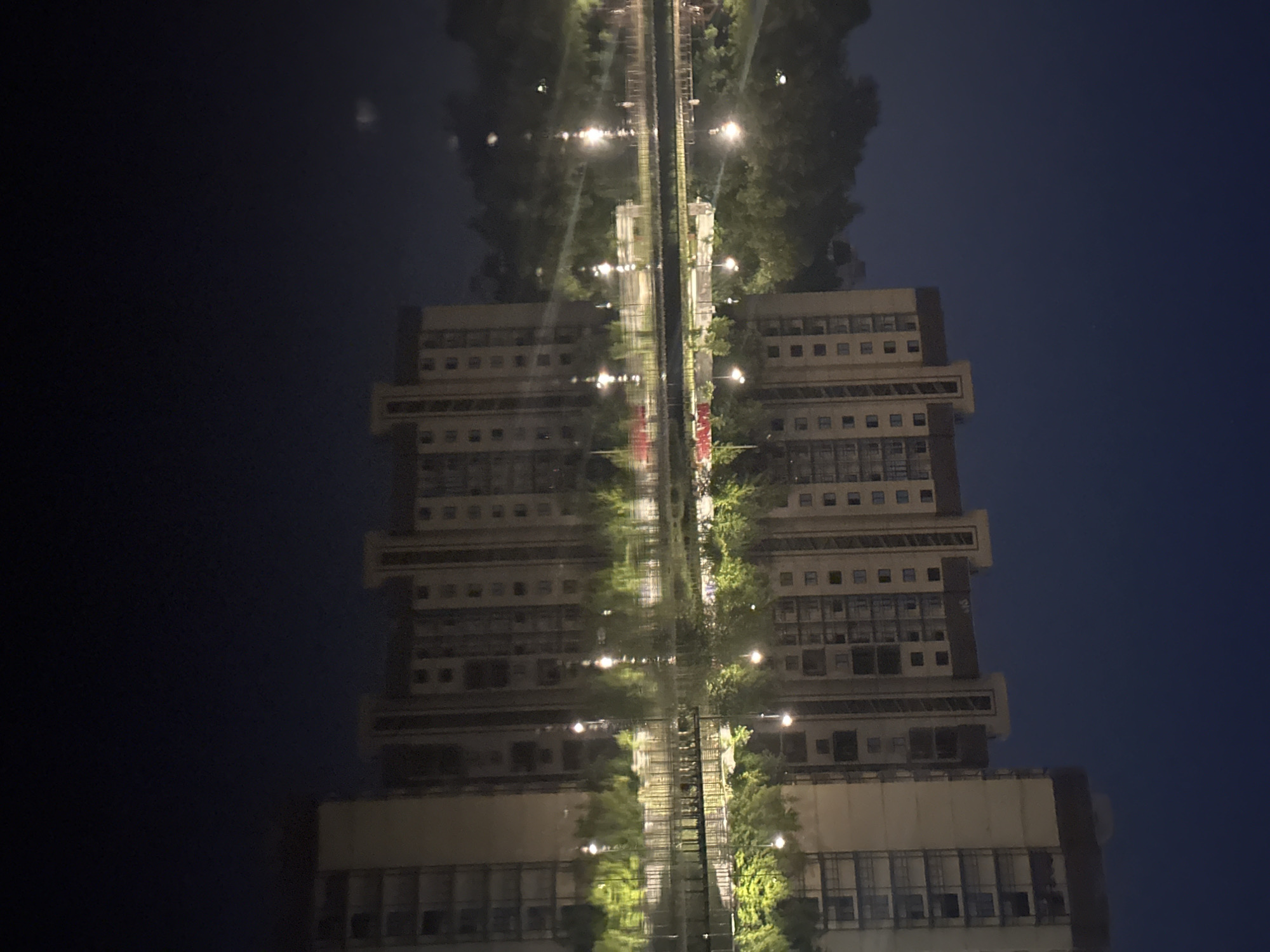 This nocturnal image depicts a tranquil waterscape in Shchyolkovo, Russia, featuring multi-story residential buildings reflected in a calm body of water. The sky, a deep navy blue, occupies the upper portion, with a singular, faint celestial object or distant light source discernible towards the upper right. On the far shore, a row of modern, light-colored apartment buildings with dark roof structures and repeating window patterns stands prominently. The building on the far right includes multiple open balconies. A dense line of deciduous trees and shrubs separates the buildings from the water's edge, intensely illuminated by numerous artificial light sources, predominantly streetlights on tall poles, casting a vivid green-yellow glow on the foliage. A wooden pathway or boardwalk is visible along the shoreline, particularly on the right, and a light-colored wall with indistinct red markings runs behind the trees on the left. The lower half of the image is dominated by the still, dark water, which perfectly mirrors the architectural structures, illuminated vegetation, and individual light sources with high fidelity, creating sharp, elongated reflections. No individuals or discernible activities are present within the scene.