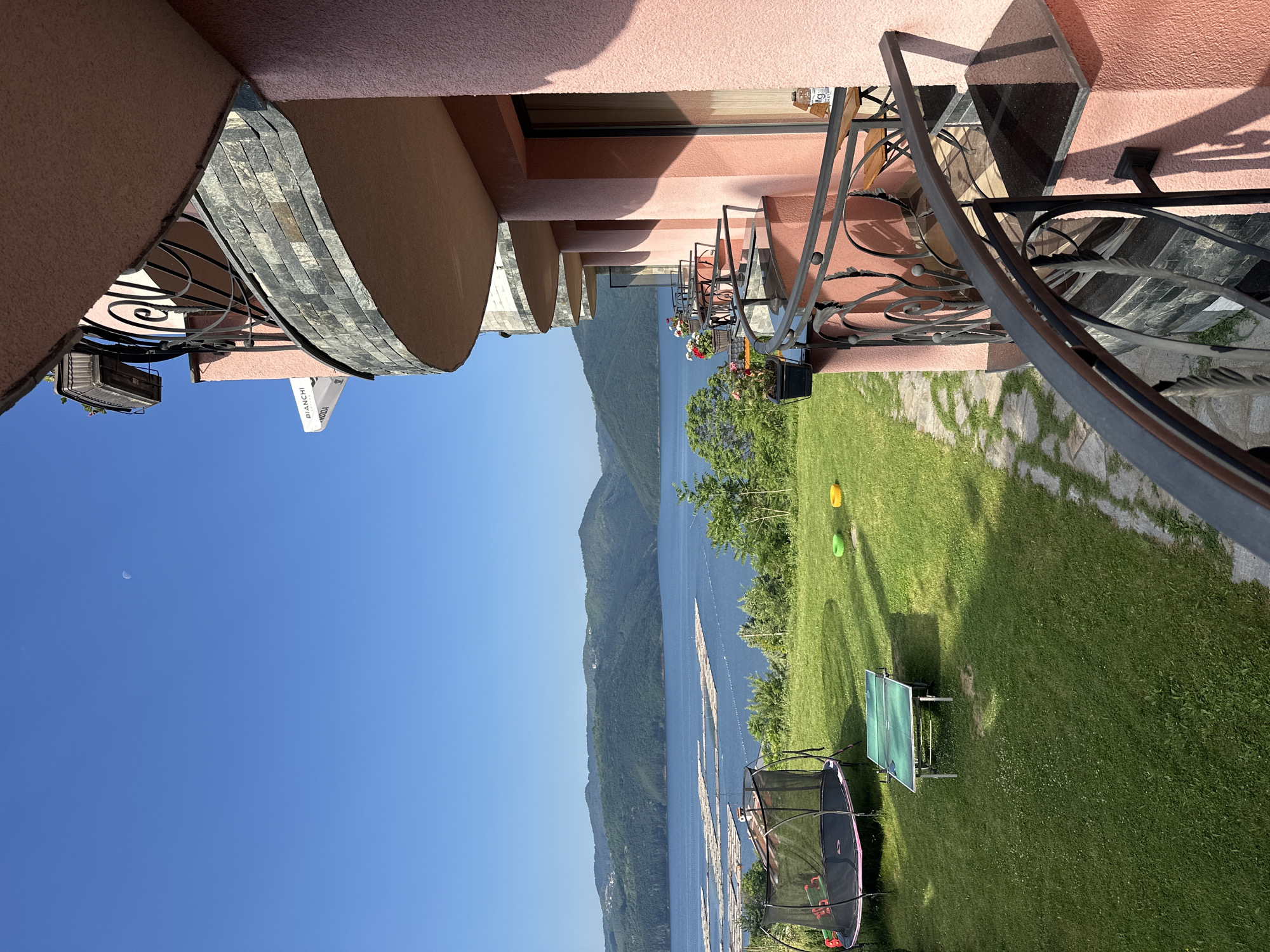 The image captures a panoramic view from a multi-story, salmon-pink stucco building with curved balconies, located in Glavatartsi, Bulgaria. The vantage point is from an upper balcony, featuring a dark ornate metal railing and a black granite-like ledge. Below, a lower balcony has a small wooden table with a bottle on it. Higher balconies, some with stone cladding and potted flowers, are visible on the right. An awning displaying "BIANCHI" and "SKODA" is partially visible.
The immediate foreground on the left features a well-maintained green lawn containing a black trampoline with a pink trim and a green ping-pong table. Two small, colored plastic objects (yellow and green) rest further back on the grass. A stone pathway delineates the right edge of the garden area.
Beyond the garden lies a large, calm body of water, likely a lake or reservoir, stretching horizontally across the middle ground. Numerous rectangular raft-like structures, possibly fish farms or logging platforms, are distributed across the water's surface. The water is bordered by forested hills and mountains, which ascend in the background. The sky above is clear and bright blue, with a faint crescent moon visible in the upper-right quadrant. The scene is brightly lit by daylight, casting shadows from the building and objects in the garden. No individuals are present in the image.