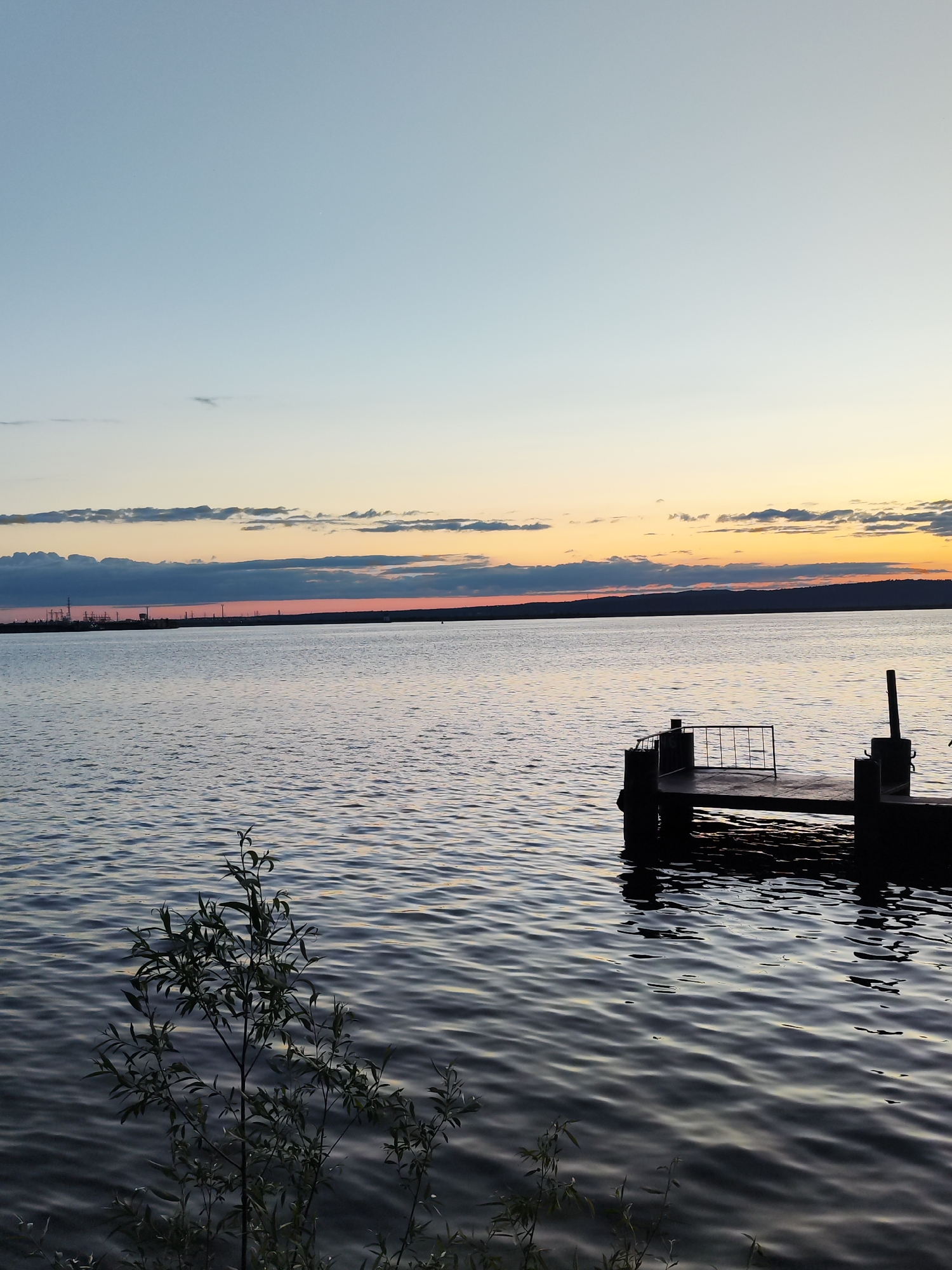 The image captures a serene twilight scene over a vast body of water, likely a large river or lake near Chaikovsky, Russia, during either sunrise or sunset. The sky, a prominent element, displays a gentle gradient, transitioning from a soft, clear blue at the top to warm hues of orange, yellow, and subtle pink closer to the horizon. Thin, elongated clouds are scattered across the lower sky, catching the light.
On the distant horizon to the left, a line of industrial structures, appearing as dark silhouettes, suggests factories or refineries, hinting at the developed nature of the area. A continuous strip of land or low hills forms the far bank, silhouetted against the colorful sky.
The body of water, possibly the Kama River, is calm with gentle ripples across its surface, faintly reflecting the muted colors of the sky. In the mid-ground on the right, a small, dark pier or dock extends into the water. It features a simple design with what appear to be metal railings, and its dark reflection is visible in the water below. To the left in the immediate foreground, a leafy bush or small tree partially frames the scene, its dark foliage providing a contrast against the water.
No people or explicit activities are visible, emphasizing the tranquility of the landscape. The scene evokes a sense of peace and quiet contemplation, marked by the soft, ambient light of either dawn or dusk. There is no visible text in the image.