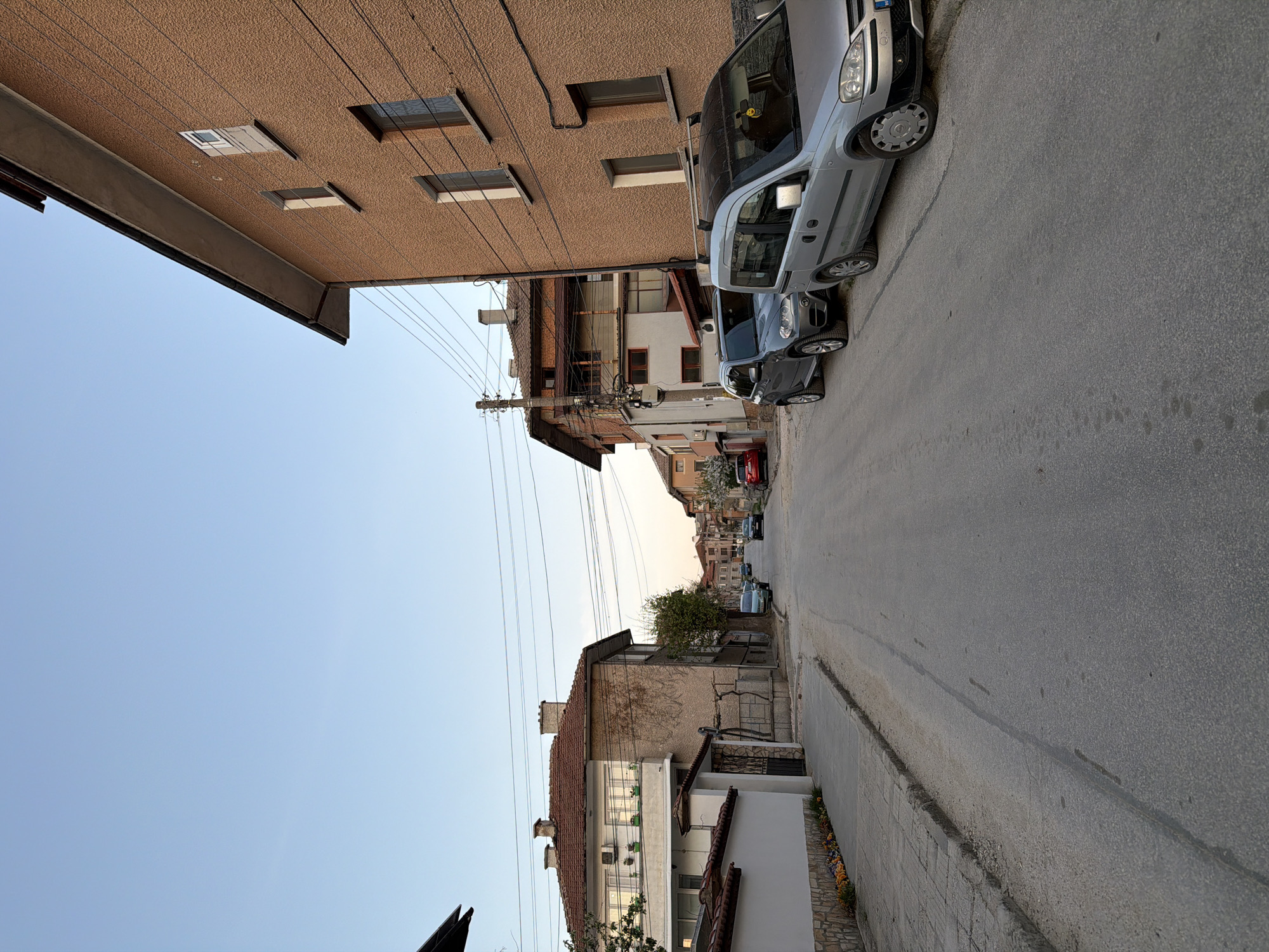 The image shows a narrow street in Panagyurishte, Bulgaria. The street is lined with buildings of varying heights and architectural styles. On the right side of the street, there are houses with tiled roofs and painted walls, while on the left, a tall, brown building with multiple windows dominates the scene. Several cars are parked along the left side of the street, including a light gray van and a dark gray car. Power lines crisscross above the street, adding to the urban landscape. The sky is a clear, pale blue, suggesting either early morning or late afternoon light. The street surface is asphalt, and a sidewalk runs along the right side.