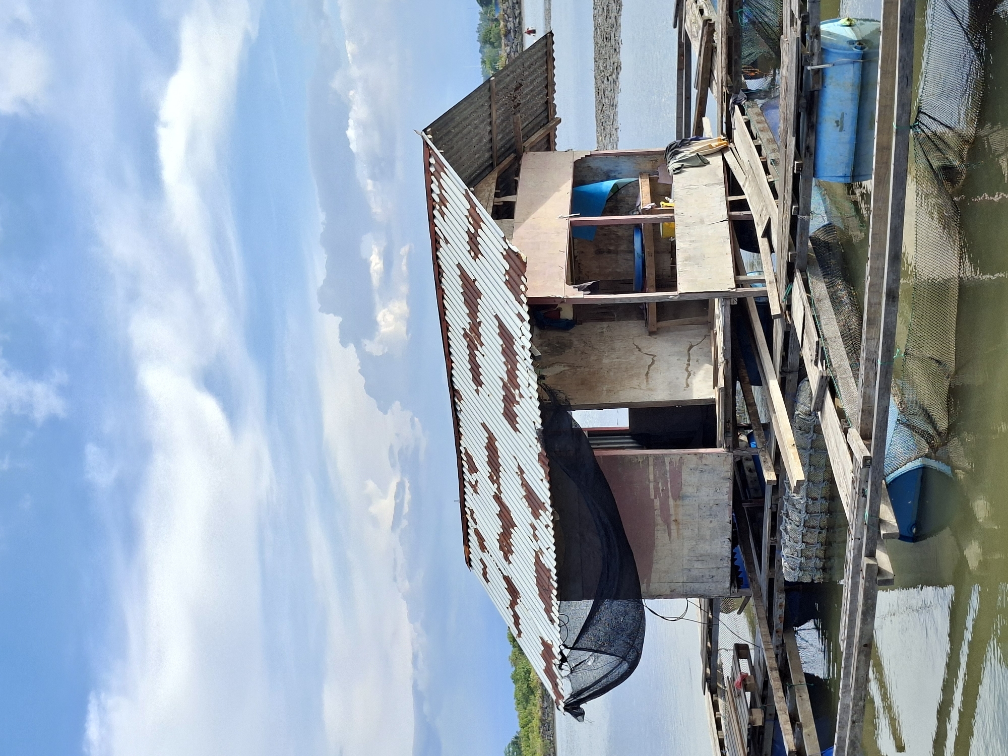 A weathered, dilapidated structure stands on stilts in a body of water under a partly cloudy sky. The structure is made of wood and corrugated metal. A large black net is attached to one side. The location appears to be Lhokseumawe, Indonesia.