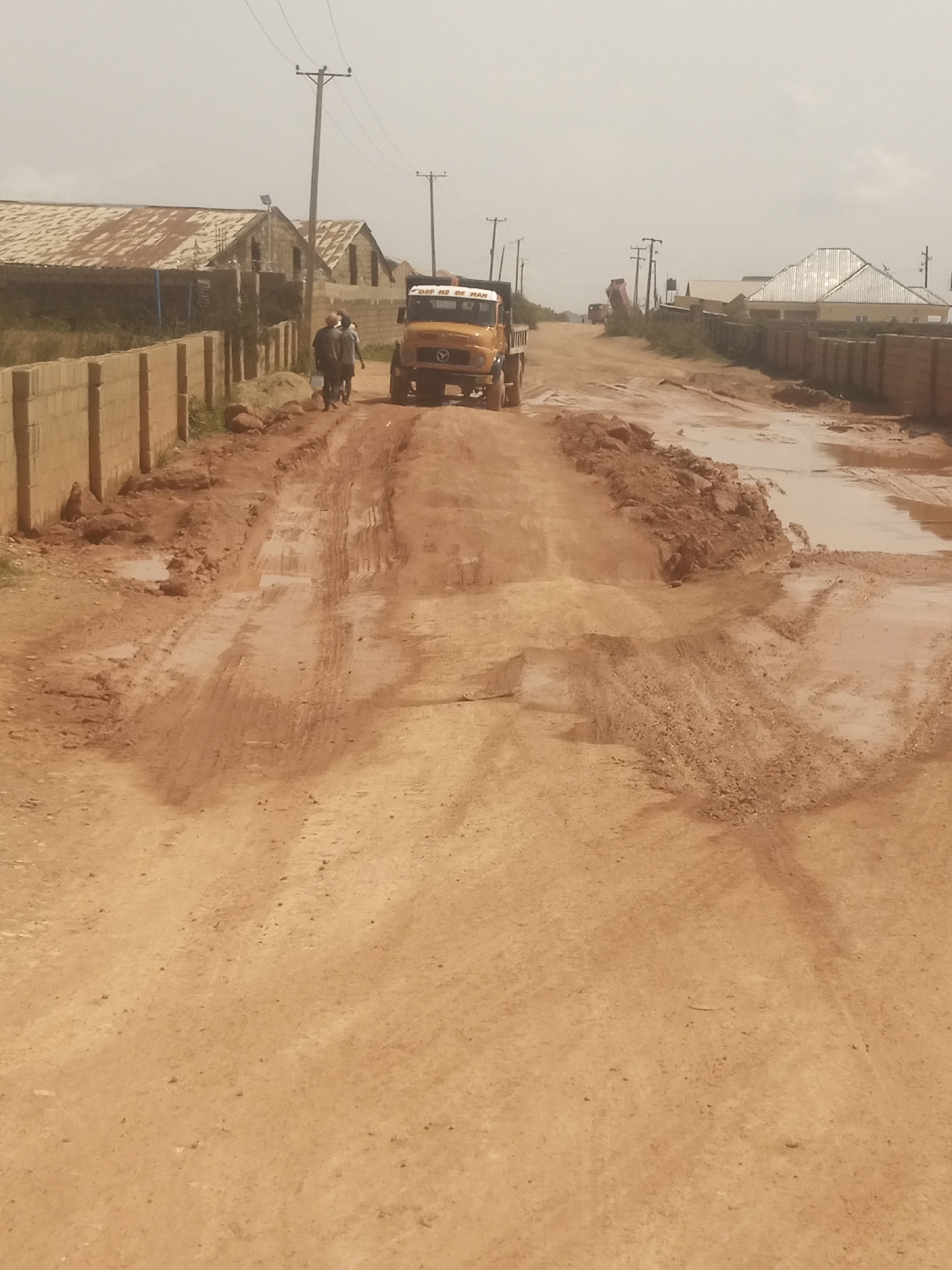 A yellow dump truck with the text "GOOD NO BE MAN" on its side is driving down a muddy, unpaved road in DU, Nigeria. Two people are walking in the distance in front of the truck. The road is wet and rutted, with puddles of water and piles of dirt. There are buildings with corrugated metal roofs and brick walls on either side of the road, as well as a low concrete fence. Utility poles with wires are visible in the background. The sky is overcast, suggesting a cloudy day. The scene appears to be a rural or developing area, possibly undergoing construction or experiencing poor road conditions due to weather.