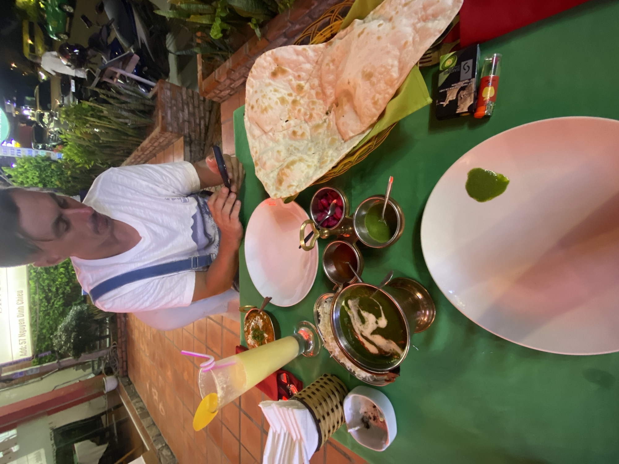 A medium shot captures a table set for a meal, likely an Indian restaurant, with a green tablecloth. In the foreground, a white plate with a dollop of green sauce is visible. To the right of this plate are a lighter and a small packet. Further back, a basket holds a large piece of naan bread. Several small bowls containing various condiments, including what appears to be a green sauce and pickled items, are arranged on the table. A glass of pale yellow liquid with two straws and a yellow garnish sits to the left, next to a napkin holder filled with white napkins. A copper bowl filled with a dark green curry is positioned behind the naan basket. Another copper bowl with a lighter colored curry is behind that. A man, wearing a white t-shirt with blue overalls, is partially visible in the upper right quadrant of the image. He is looking down at his phone, which he holds in his left hand. The background suggests an outdoor seating area of a restaurant, with potted plants and illuminated signage indicating "Add: 57 Nguyen Dinh Chieu," a street in Phu Thuy Ward, Vietnam. Motorcycles are parked in the background, and a yellow van is visible further down the street. The lighting suggests it is evening.