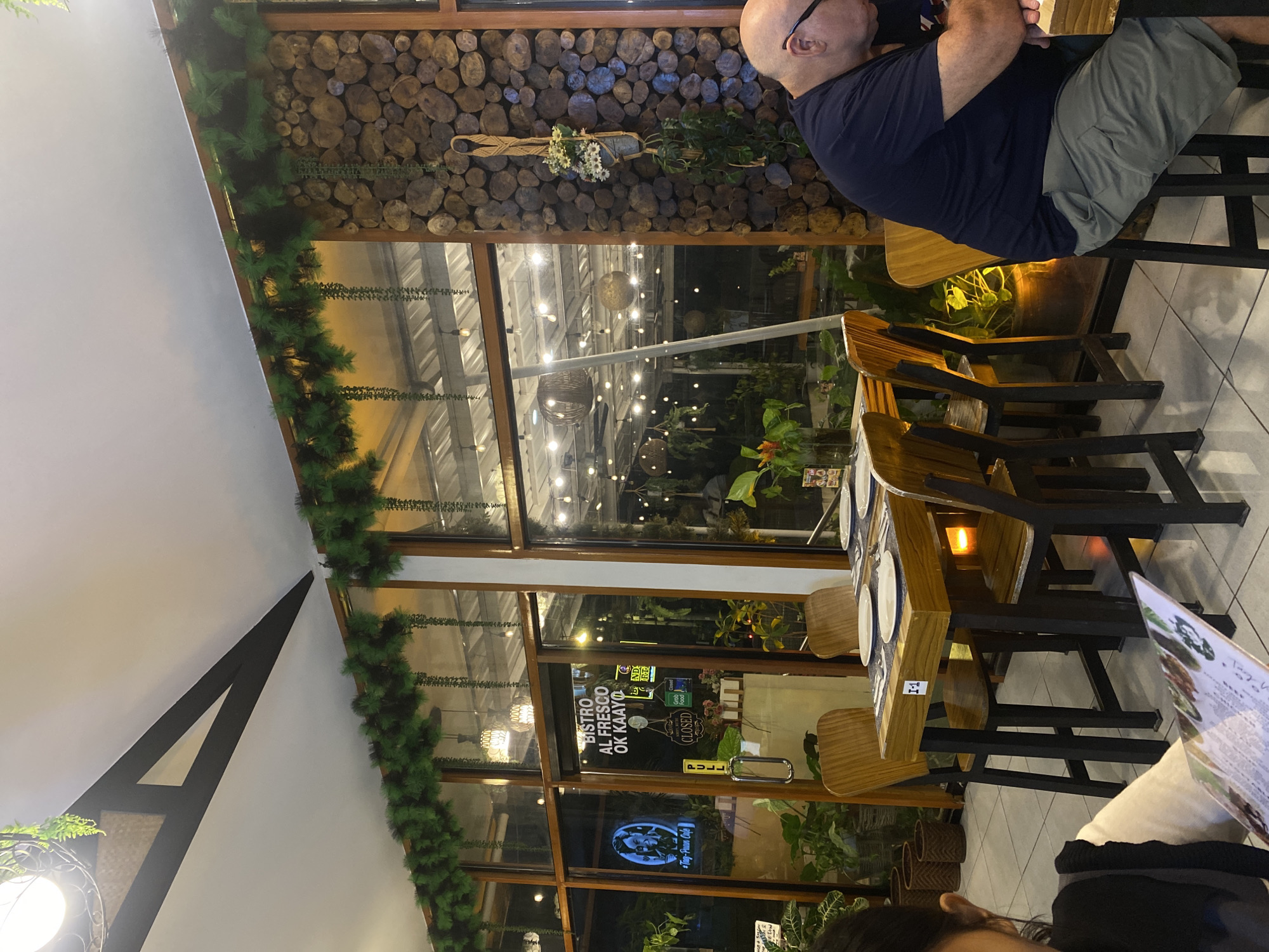 The image shows the interior of a restaurant in Davao City, Philippines. The restaurant has a rustic and natural ambiance, with wooden furniture, decorative logs on the wall, and abundant greenery. There are tables set with plates and cutlery, and chairs arranged around them. Through the large glass windows, the outdoor area of the restaurant is visible, illuminated by string lights. In the foreground, a person is seated at a table, holding a menu. Another person is partially visible in the bottom left corner of the image. The lighting is warm and inviting, creating a cozy atmosphere. The restaurant appears to be decorated with festive garlands, suggesting a holiday season.