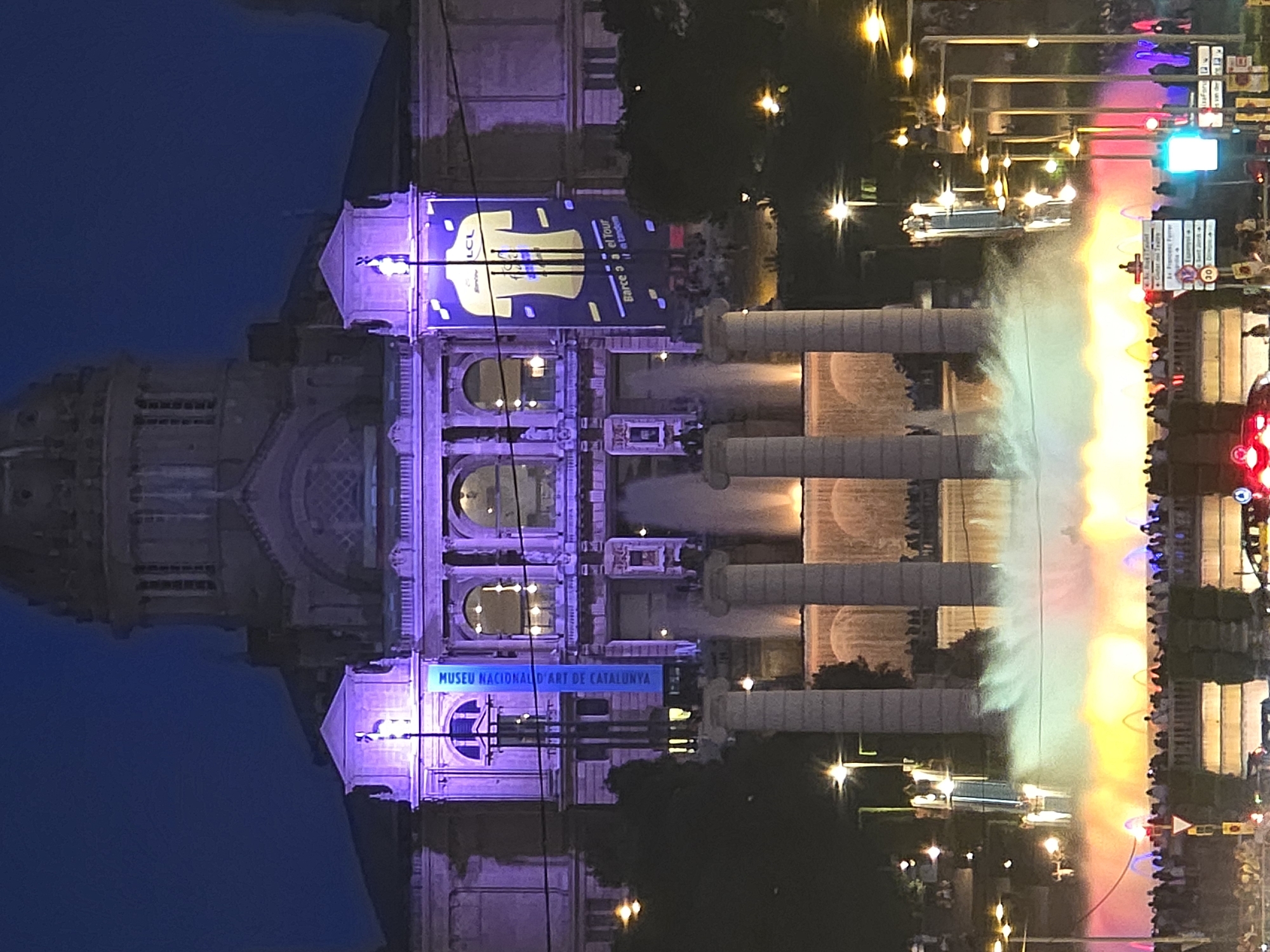 The image depicts a night scene at the Magic Fountain of Montjuïc in Barcelona, Spain. A grand, illuminated building, identified by a banner reading "MUSEU NACIONAL D'ART DE CATALUNYA," serves as the backdrop. The building is lit with purple lights, and banners advertising the "LCL" and "Barce a el Tour" are displayed. In the foreground, the Magic Fountain is in action, with water jets creating a dynamic display of colorful lights and mist. People are gathered on a bridge and in front of the fountain, observing the spectacle. Streetlights and traffic lights are also visible, indicating an urban environment. The sky is dark, suggesting it is nighttime.