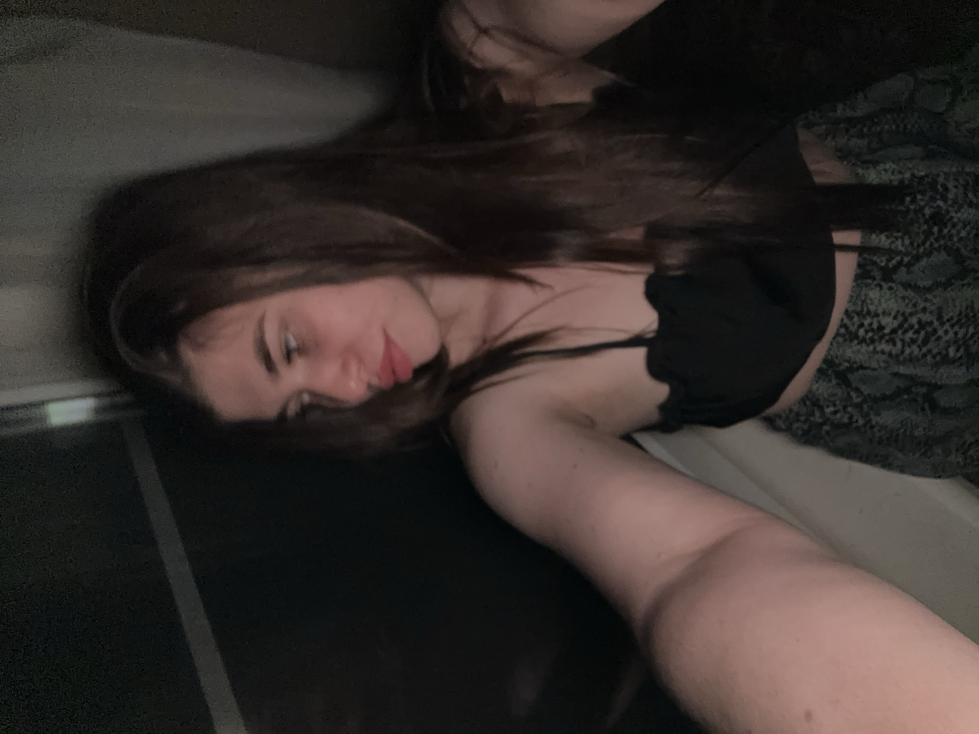 A young woman with long brown hair is reclining on what appears to be a bed or cushioned surface. She is wearing a black top with spaghetti straps and a grey patterned skirt or pants. The lighting is dim. The background is blurry, but the presence of a metallic edge suggests the woman is next to a window. There is no evidence in the image to suggest the location is in the city of Мещёры, Russia.