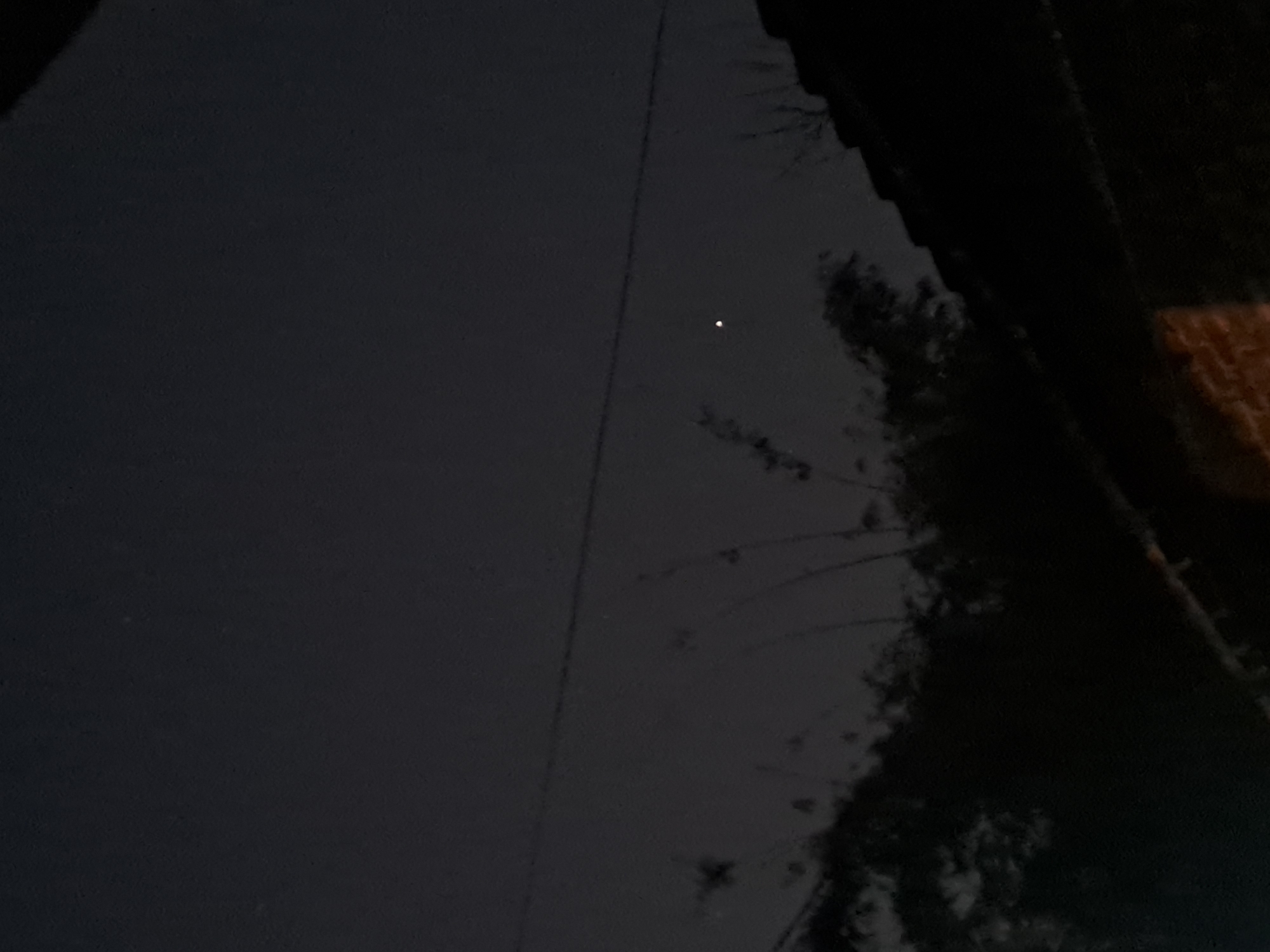 The image shows a night sky with a single bright celestial object, likely a planet or a very bright star, visible above silhouetted trees and power lines. The lower portion of the image is dominated by the dark roof of a building. The location in Sumberejo, Indonesia, is not directly ascertainable from the visual information provided in the image. There are no people or animals present, nor any observable actions or interactions. The environment is dark, indicating nighttime, with a clear or mostly clear sky. The overall scene is still and quiet.