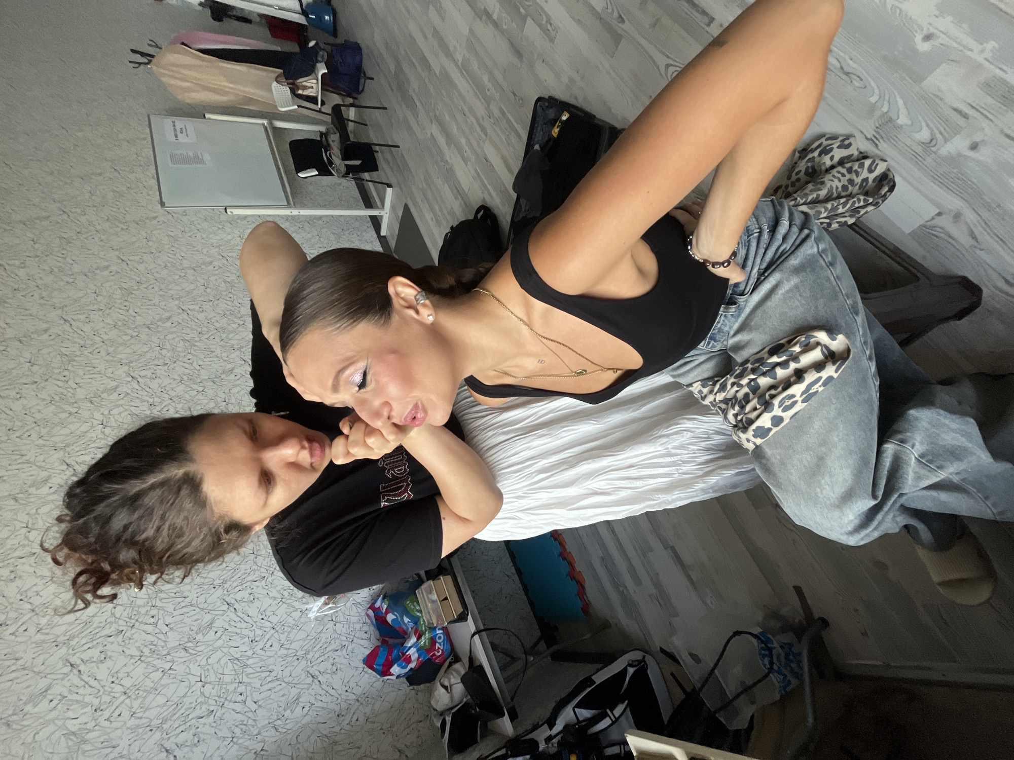 In a brightly lit room, a woman with dark, curly hair pulled up high is applying makeup to another woman. The makeup artist wears a black t-shirt and a long, white, pleated skirt. The subject, seated on a stool, wears a black tank top and light-wash jeans. Her hair is pulled back in a sleek bun, and she has dramatic makeup on her eyes, including glittery eyeshadow. She is also wearing gold necklaces and large hoop earrings. A leopard-print scarf is tucked into her waistband.
The room appears to be a studio or backstage area. To the left, a table is cluttered with various items, including bags, boxes, and what looks like a makeup kit. In the background, a whiteboard stands on a stand, with papers attached. Several chairs are visible, along with clothes hanging on racks and bags on the floor. The floor is light-colored wood. The overall impression is one of preparation for a performance or photoshoot. The image does not contain information that allows for determining the city of Moscow, Russia.