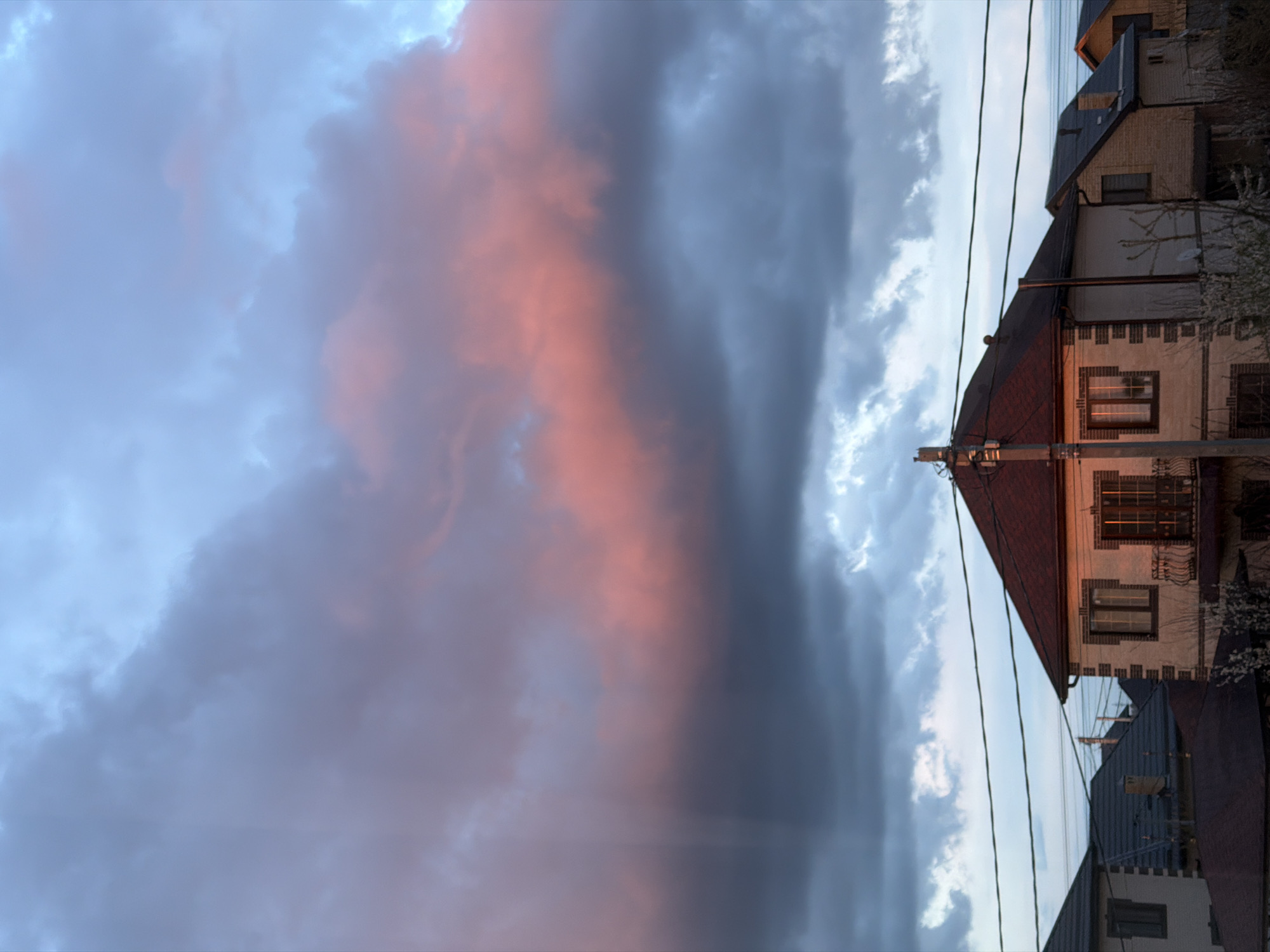 The image depicts a residential area, possibly Rossiyskiy, Russia, under a dramatic sky with a mix of dark and light clouds illuminated by the setting sun, casting a pinkish hue. Several multi-story houses are visible, featuring varied architectural styles. Power lines traverse the scene in the foreground. No people are visible.