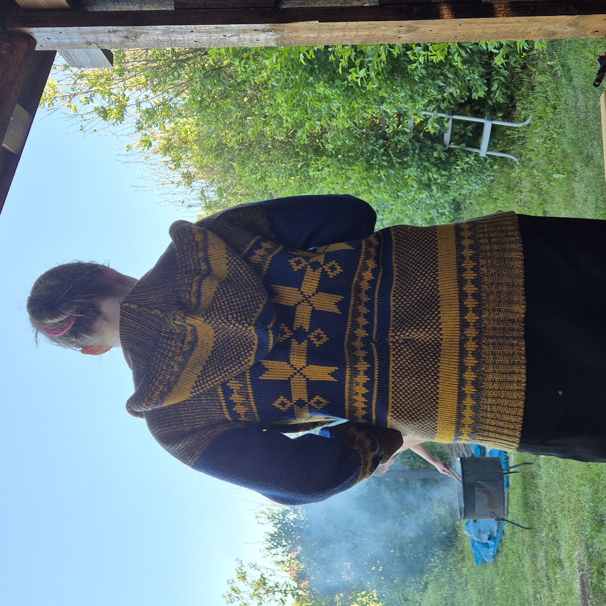 A person, viewed from the back and slightly from above, is wearing a hooded, knitted sweater with a dark blue and yellow patterned design. The sweater has a prominent geometric pattern on the back, featuring stylized crosses and diamond shapes. The person has their hair pulled back with a pink accessory. They appear to be outdoors, possibly in a backyard or a rural setting, with lush green trees and grass visible in the background. The sky is a clear blue, suggesting a sunny day. In the distance, a small, dark-colored barbecue grill is visible, with a wispy trail of smoke rising from it. The person's left hand is extended towards the grill, holding a metal utensil, indicating they might be cooking or tending to the grill. The overall scene suggests a casual, outdoor activity. The time of day appears to be daytime, likely late morning or afternoon, given the bright sunlight and clear sky. There is no visible text in the image. The setting could be a "Novostroyka" (new construction) area in Russia, which often features newly built houses with surrounding undeveloped land or gardens.