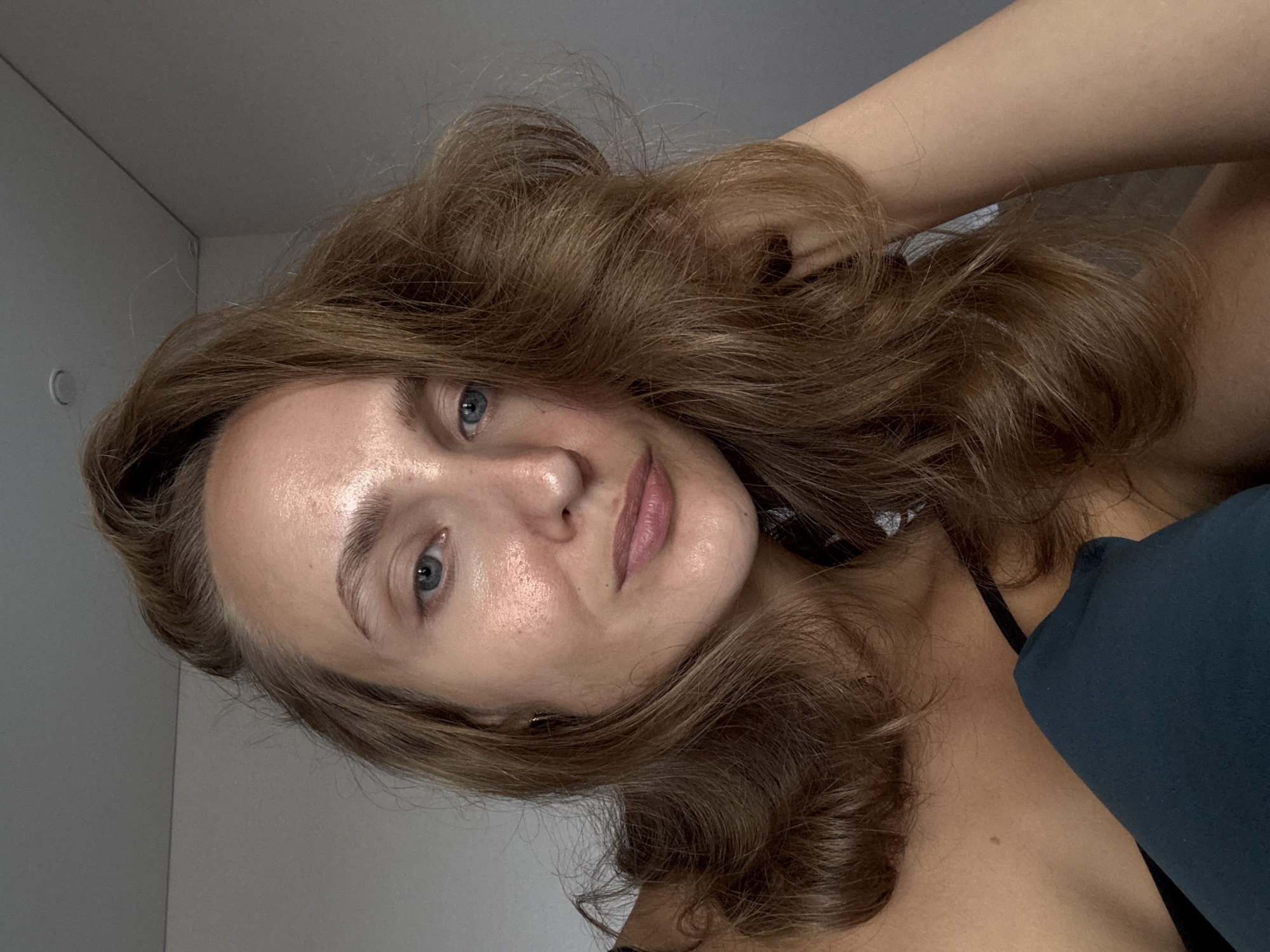 A young woman with long, wavy, reddish-brown hair is positioned in the foreground, facing the camera. Her head is tilted slightly, and her right arm is resting on the back of her head. She has light skin with a dewy complexion, blue eyes, and light brown eyebrows. Her lips are slightly parted, and she is wearing a black camisole. A dark teal pillow or cushion is visible in the lower left quadrant of the image, partially obscuring her chest and shoulder. The background consists of a plain, light-colored wall that transitions to a darker gray color in the upper right corner, suggesting an indoor setting. There are no discernible elements that place the scene in any specific city or geographical location.