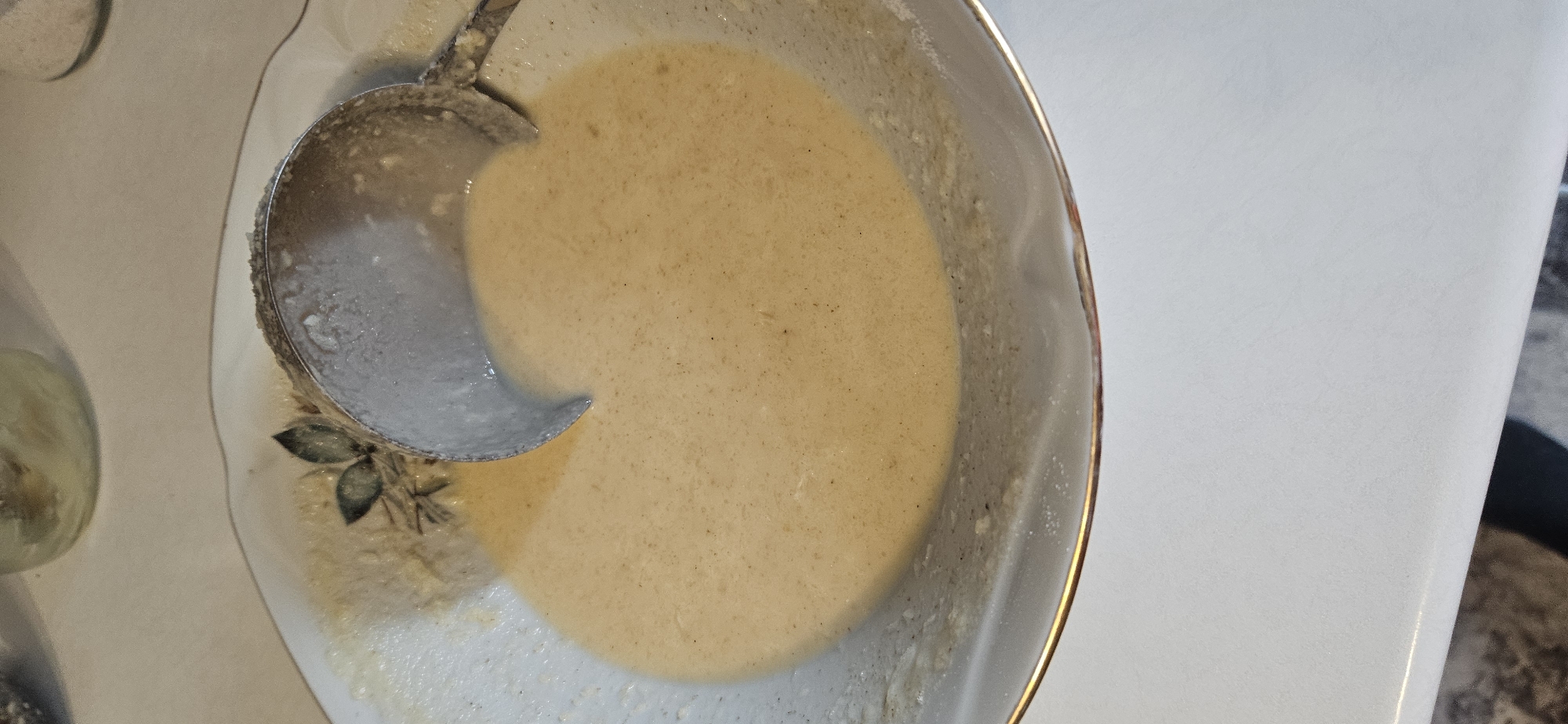A close-up, overhead shot captures a shallow, decorative bowl filled with a pale, beige batter, likely for pancakes or crepes. A metal ladle, also coated in the batter, rests within the bowl, its handle extending out of frame to the right. The bowl's rim features a subtle gold trim and a faded floral pattern. The background is a plain, light-colored surface, possibly a table. No specific location in Vitsebsk, Belarus, can be identified from the image alone. The scene depicts a food preparation moment.