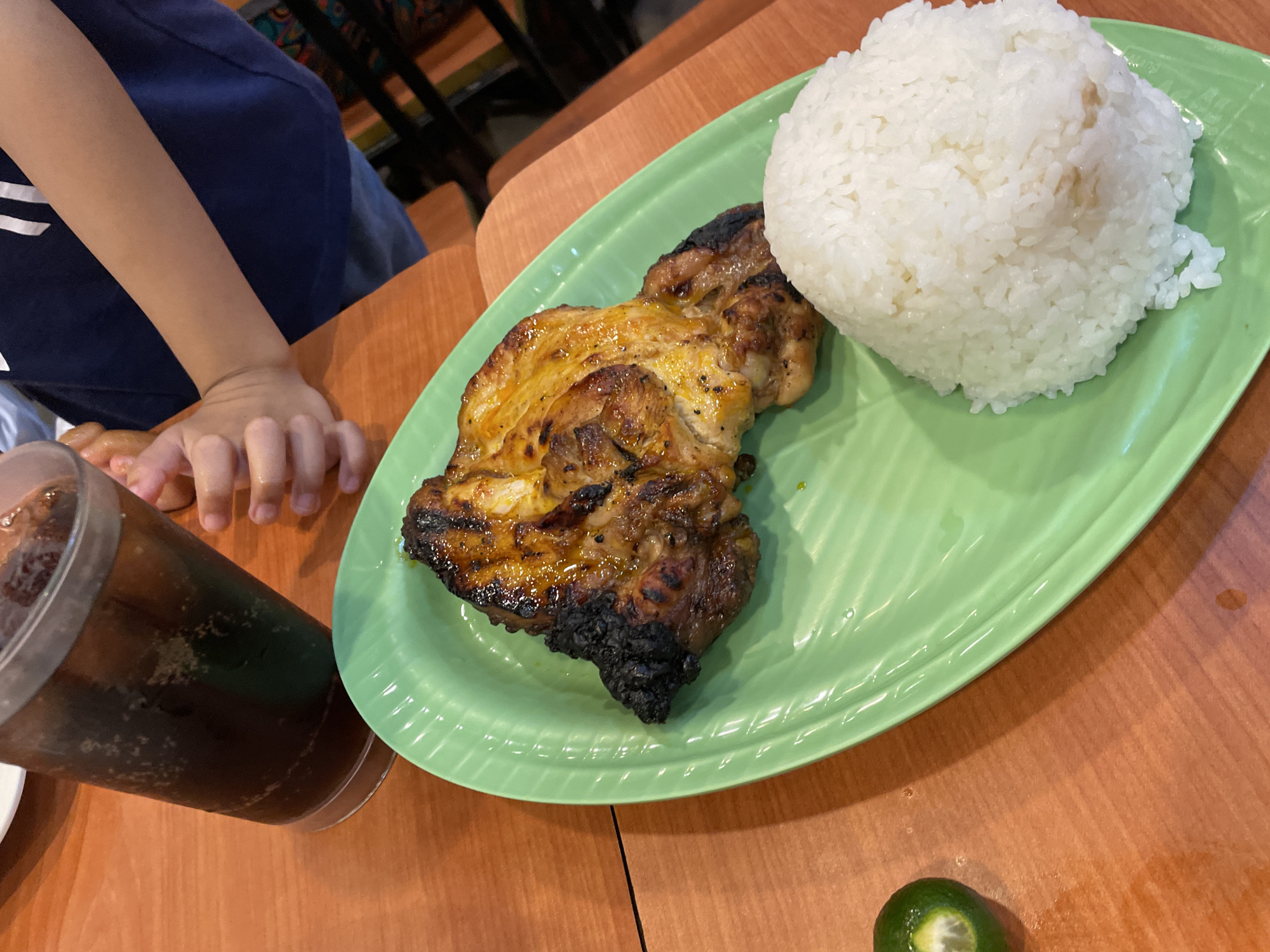 The image depicts a meal served on a light brown wooden table. The central subject is a bright green, oval-shaped plate with a textured, ribbed surface, holding a portion of grilled chicken and a mound of white rice. The grilled chicken, likely a thigh and leg piece, exhibits a glossy, orange-brown exterior with some charred sections, indicative of a marinade or basting sauce. To the left of the chicken, a quarter of a small green citrus fruit, appearing to be calamansi, is visible on the table. In the upper left quadrant, a clear plastic cup containing a dark, carbonated beverage with ice and visible bubbles is positioned. In the upper right quadrant, the forearms and hands of a child are visible, wearing a navy blue t-shirt with white vertical stripes on the left shoulder area. The background elements are out of focus but suggest an indoor restaurant setting with dark and possibly colorful furniture or decor. Based on the distinctive green ribbed plate and the type of grilled chicken (resembling *chicken inasal*) served with rice and calamansi, the scene is consistent with a **Mang Inasal** restaurant location in Davao City, Philippines.