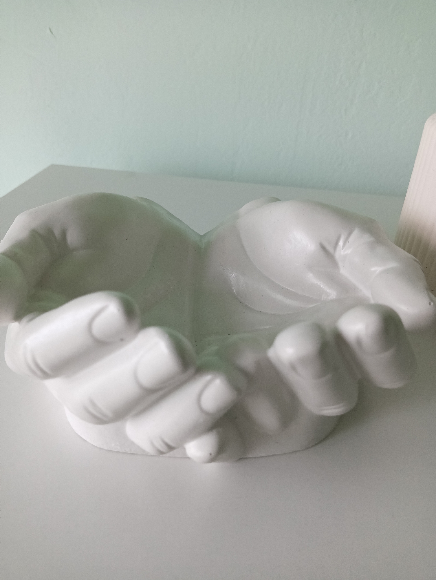 A white, ceramic decorative bowl shaped like cupped hands sits on a white surface against a light green wall. The bowl is hollow in the center, with the fingers and palms of both hands forming its structure. The image is a close-up shot, focusing on the bowl and its immediate surroundings. No people are present in the image. The location is the city of Nizhnetroitskiy, Russia.