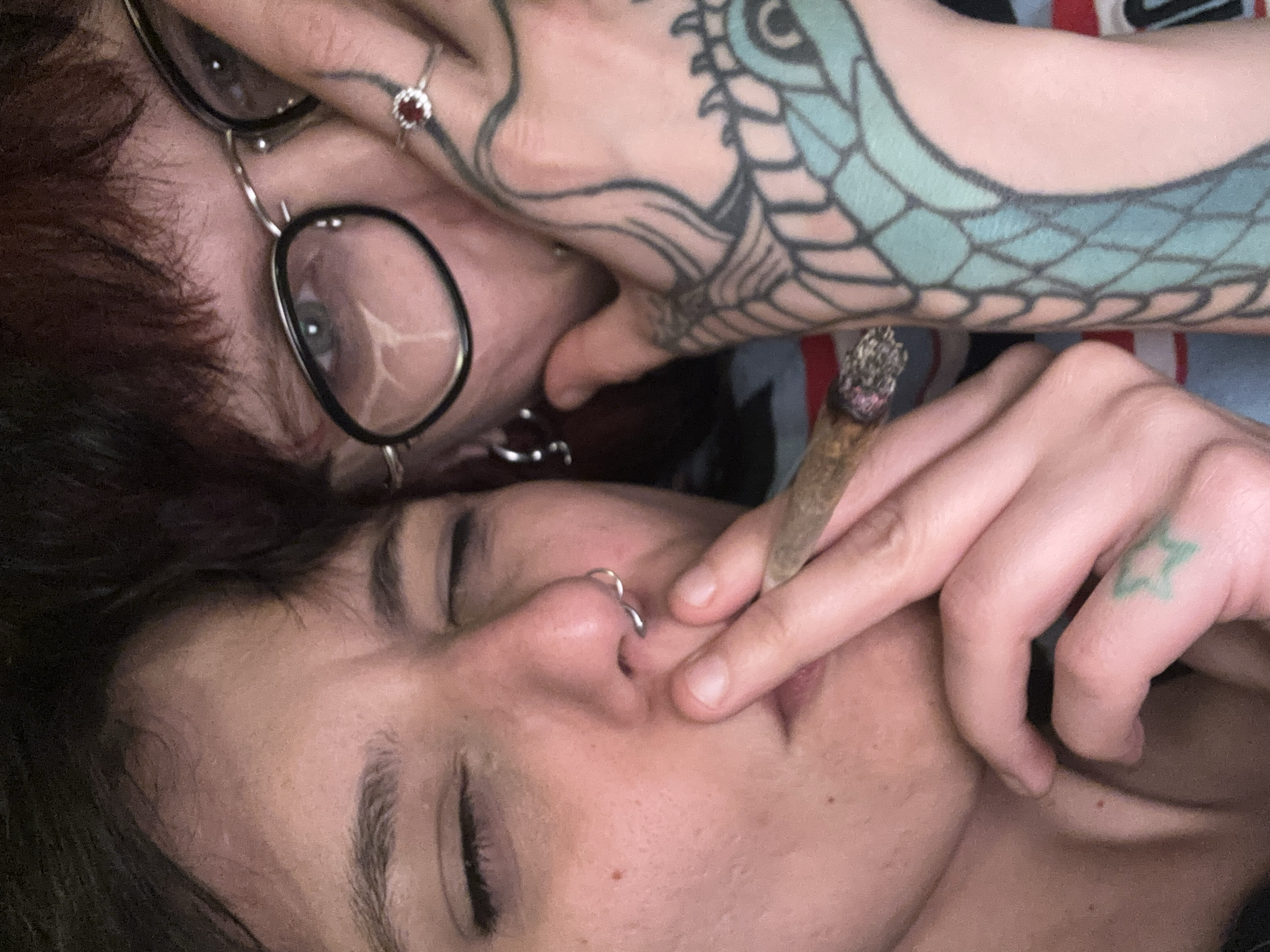 Two individuals are lying down, close together. One person, wearing round, black-rimmed glasses and a ring with a red stone, is partially visible with their eye open. The other person has their eyes closed, a nose ring, and is holding a lit cigarette or joint between their fingers. Tattoos are visible on both individuals' hands and arms, including a dragon design and a green star. The background appears to be a patterned fabric. There is no verifiable information in the image to indicate that it was taken in Sofia, Bulgaria.