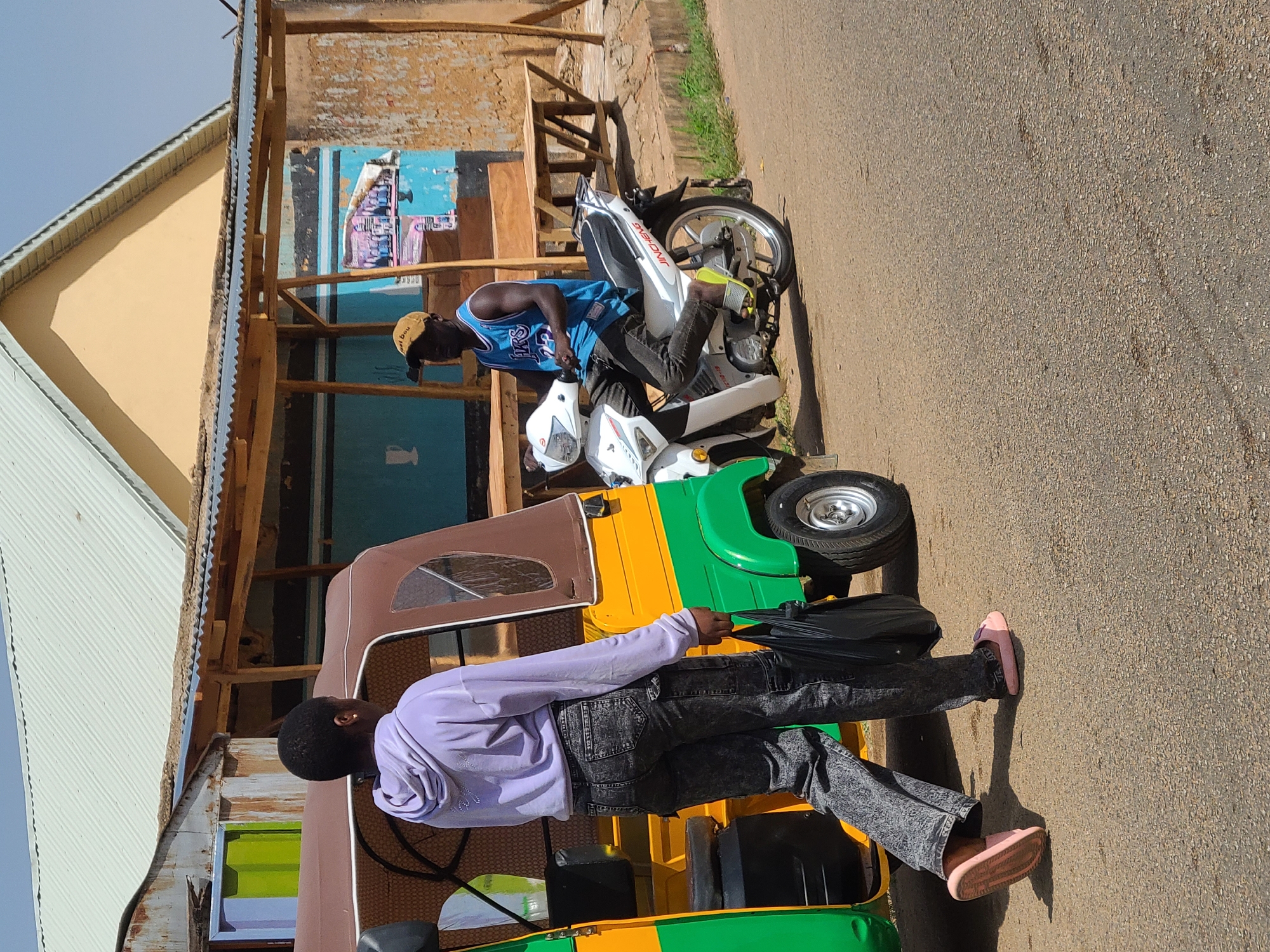 A medium, angled shot shows two African men with a tuk-tuk (auto rickshaw) and motorbike in Jos, Nigeria. The man near the tuk-tuk, wearing a long-sleeved light purple shirt, acid-wash jeans, and pink sandals, walks away from the vehicle. The other man sits on a white motorbike in a blue jersey and cap. The vehicles are on a paved surface near buildings with corrugated metal roofs and a wooden structure.