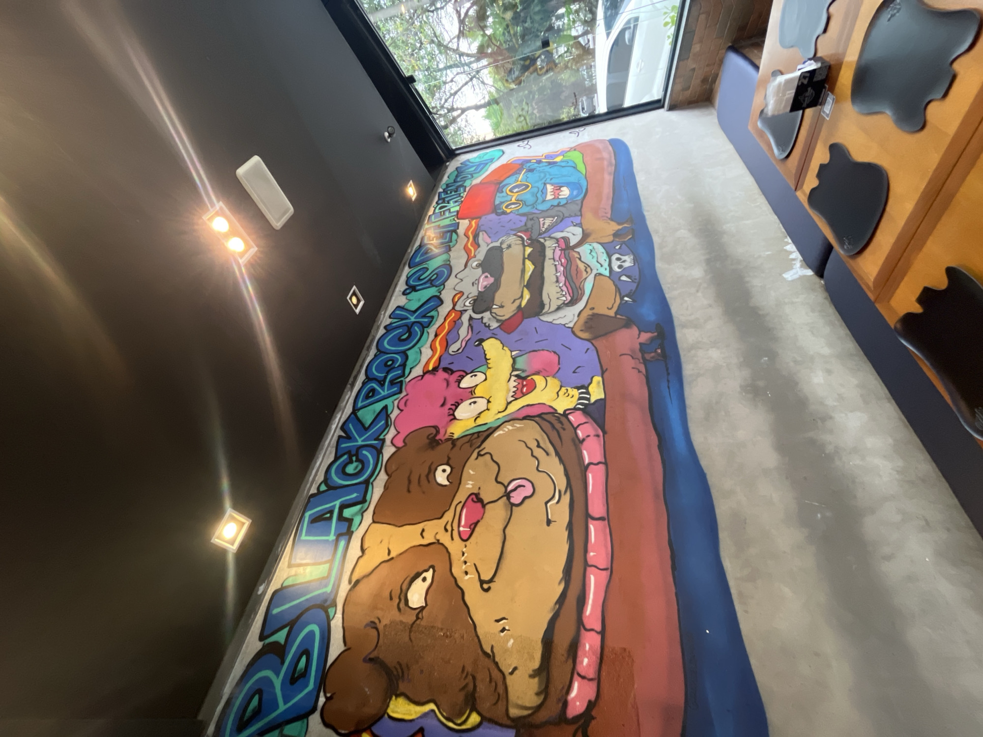 The image shows a mural inside a restaurant in an unknown city. The mural features cartoon characters with graffiti-style lettering. It's painted on the floor along the wall, near a window and tables with cow-shaped placemats. The restaurant has dark walls with recessed lighting. The scene appears to be indoors and well-lit.