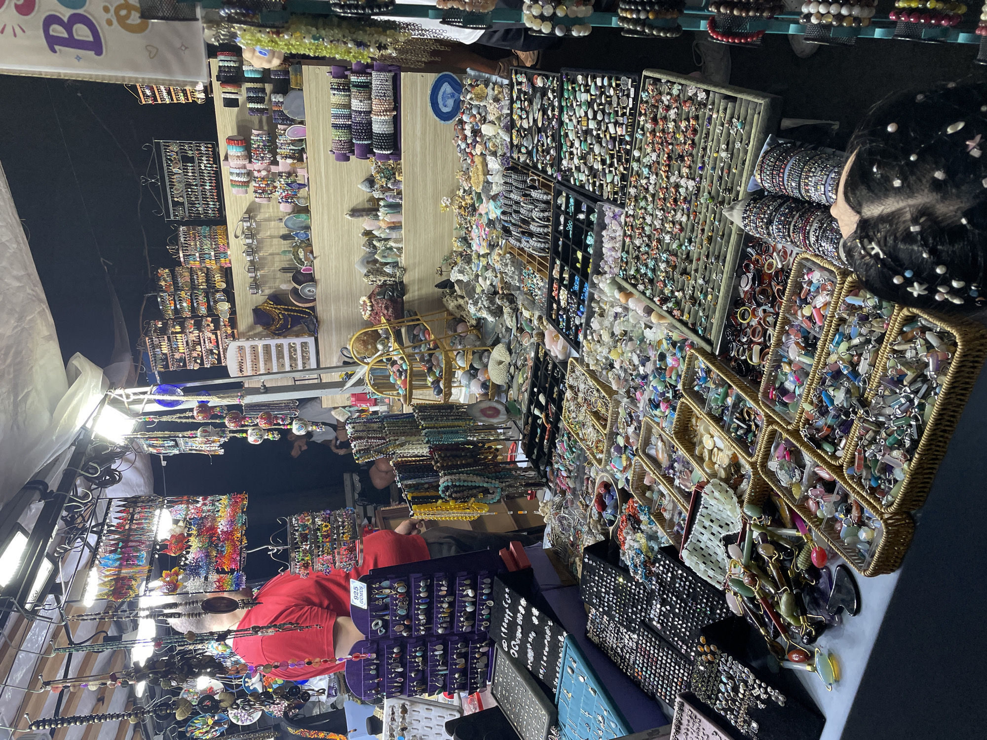 The image depicts a bustling outdoor market stall at night, likely in Kadıköy, Turkey, given the context. The stall is laden with a vast array of jewelry and decorative items, primarily made of semi-precious stones, beads, and metal. In the foreground, a person's head is visible, with decorative hair clips and pearls adorning their dark hair. They are looking down at the merchandise.
The stall is organized with various displays. On the left, there are racks and trays filled with necklaces, bracelets, and pendants. A man wearing a red t-shirt is partially visible behind some of these displays, appearing to be an vendor or shopper. Further back, shelves are stocked with more jewelry, including earrings and necklaces hung on display stands. In the center and right of the image, numerous shallow baskets and trays overflow with rings, bracelets, and loose stones, showcasing a wide spectrum of colors and textures. A prominent display features numerous stone rollers and gua sha tools.
Overhead, artificial lights illuminate the scene, casting a warm glow on the merchandise. Tarpaulins or awnings are visible above the stall, suggesting an outdoor setting. The overall impression is one of vibrant commerce, with a rich assortment of handcrafted goods presented for sale. The atmosphere appears lively and engaging.