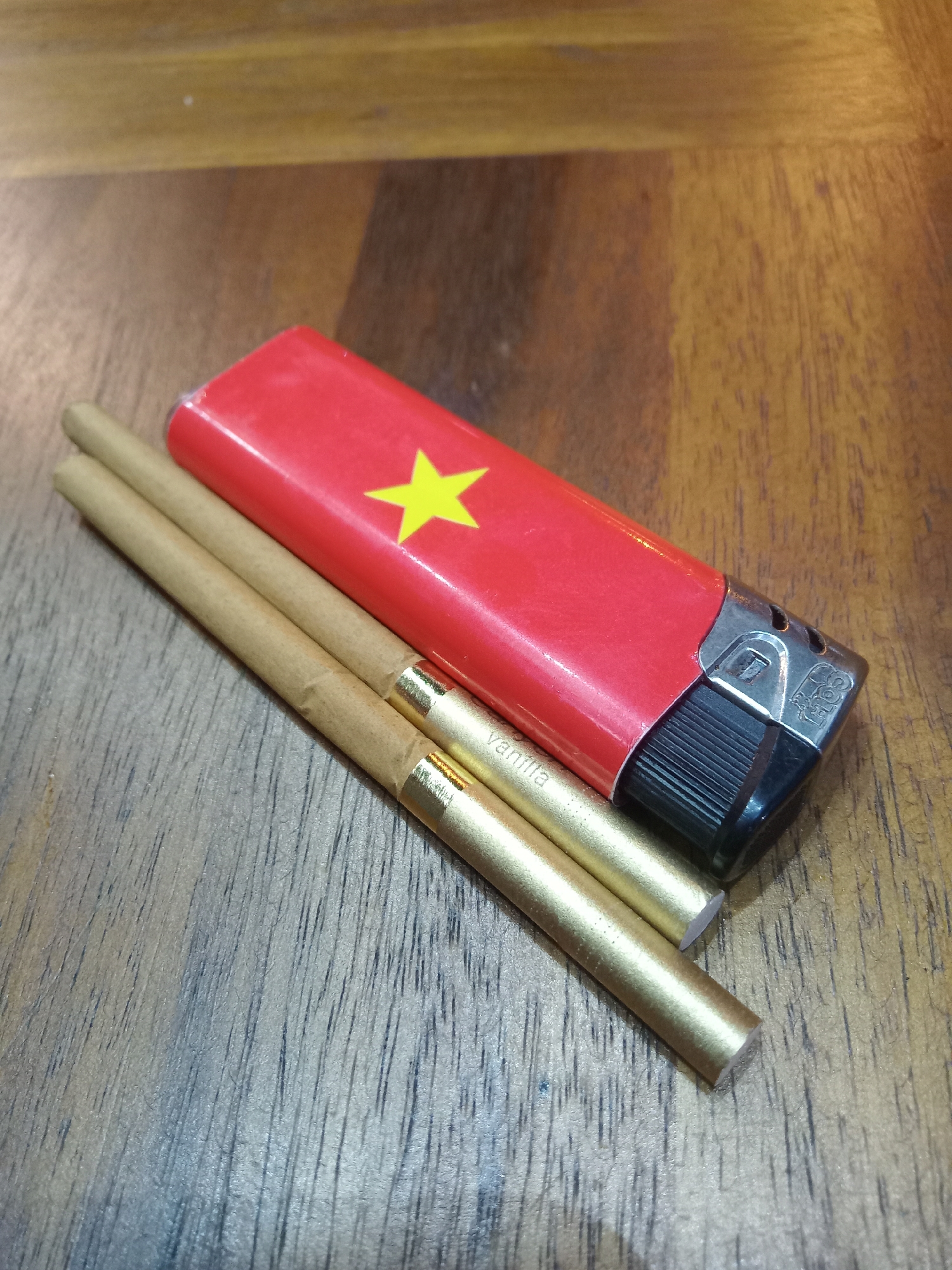 On a wooden surface, likely a table, are three slender, cylindrical objects that appear to be vanilla flavored cigars. They are positioned parallel to each other. Adjacent to the cigars is a red lighter with a yellow star, resembling the flag of Vietnam. The lighter has a black ignition mechanism and a silver-colored top. Given the Vietnamese flag depicted on the lighter, the image was probably taken in Vietnam. It is not possible to determine if this photograph was taken in Nha Trang specifically without further information.
