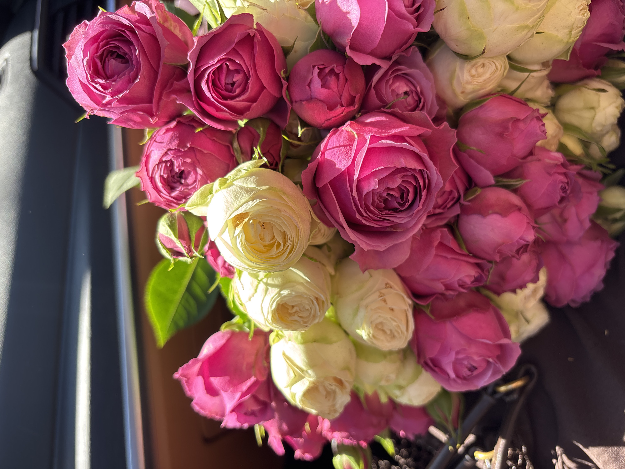 The image shows a bouquet of pink and white roses. The roses are tightly bunched together, with the pink roses predominating. The white roses are interspersed throughout the bouquet. The bouquet appears to be sitting on a dark surface, possibly a car seat, with a partially visible car window to the left. The lighting is bright and sunny. The location in Yerevan, Armenia is not discernible from the image.