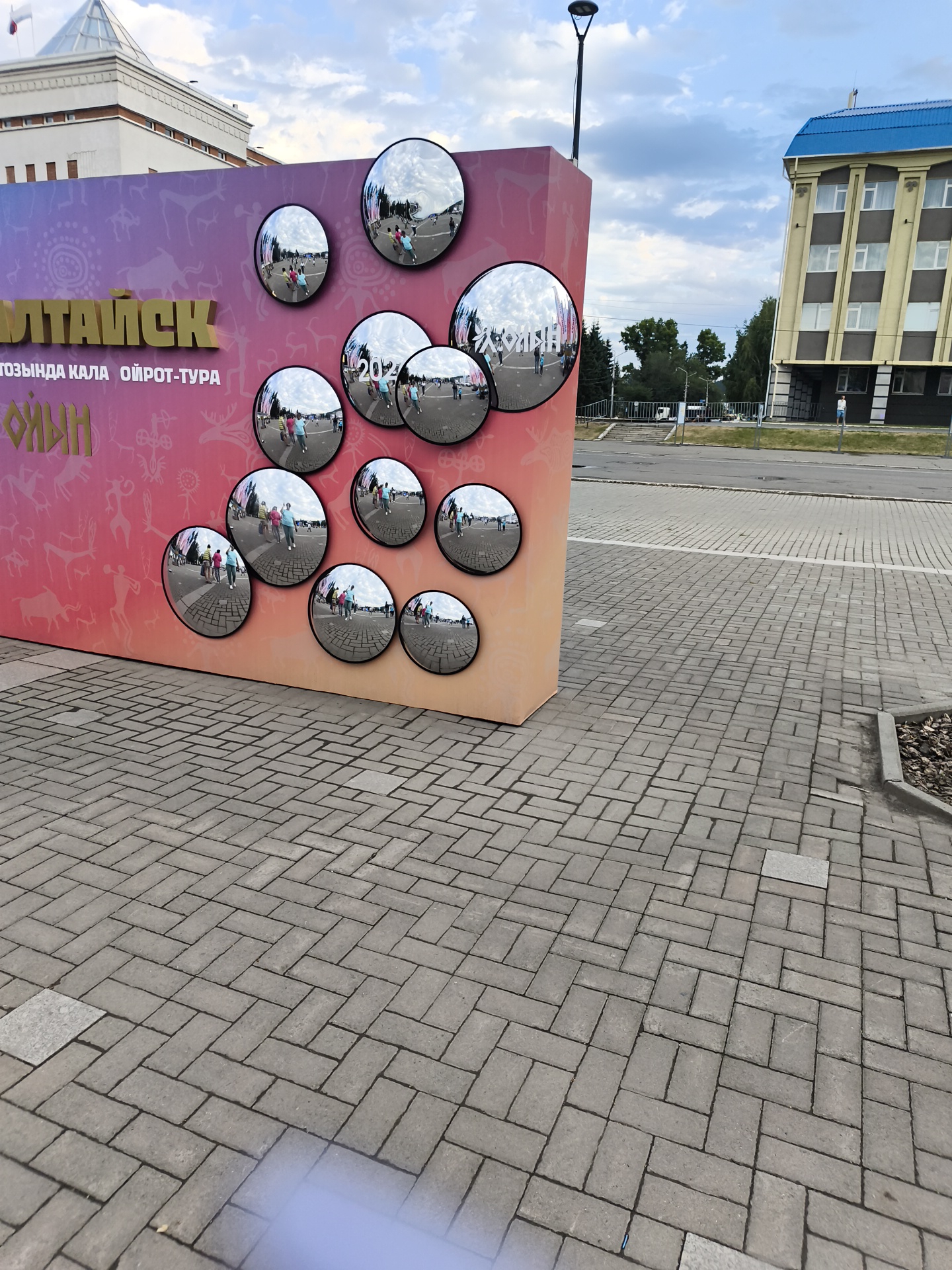 In Gorno-Altaysk, Russia, a large, colorful sign featuring text and decorative elements is displayed outdoors. The sign, which appears to be part of an informational or artistic installation, has a gradient background transitioning from pink to orange. Several circular, convex mirrors are attached to the surface of the sign, reflecting the surrounding environment. These mirrors capture images of people walking on a paved area, as well as buildings and the sky. The ground in front of the sign is paved with interlocking gray bricks laid in a herringbone pattern. In the background, a modern building with a blue roof and a lighter facade is visible, along with trees and a street. The sky is partly cloudy.