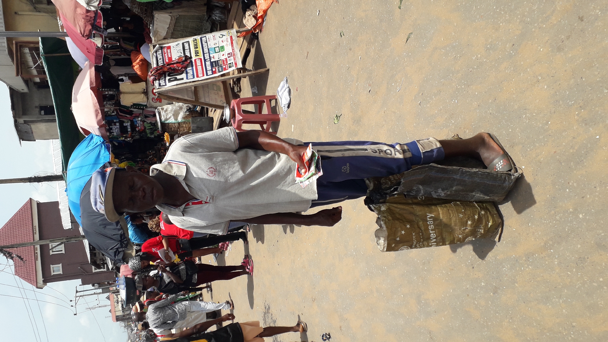 The image depicts a street scene in Onitsha, Nigeria. A middle-aged man is the central subject, wearing a light-colored shirt, dark blue sweatpants, and a hat. He is standing on two bags filled with stuff with one foot on each bag, holding a stack of small paper items, presumably money. Behind him, there are umbrellas, a small red stool, and a sign advertising money deposit and withdrawal services, bill payment options, and airtime/data subscriptions. Other people are in the background, and surrounding buildings are also visible. The ground appears to be paved.