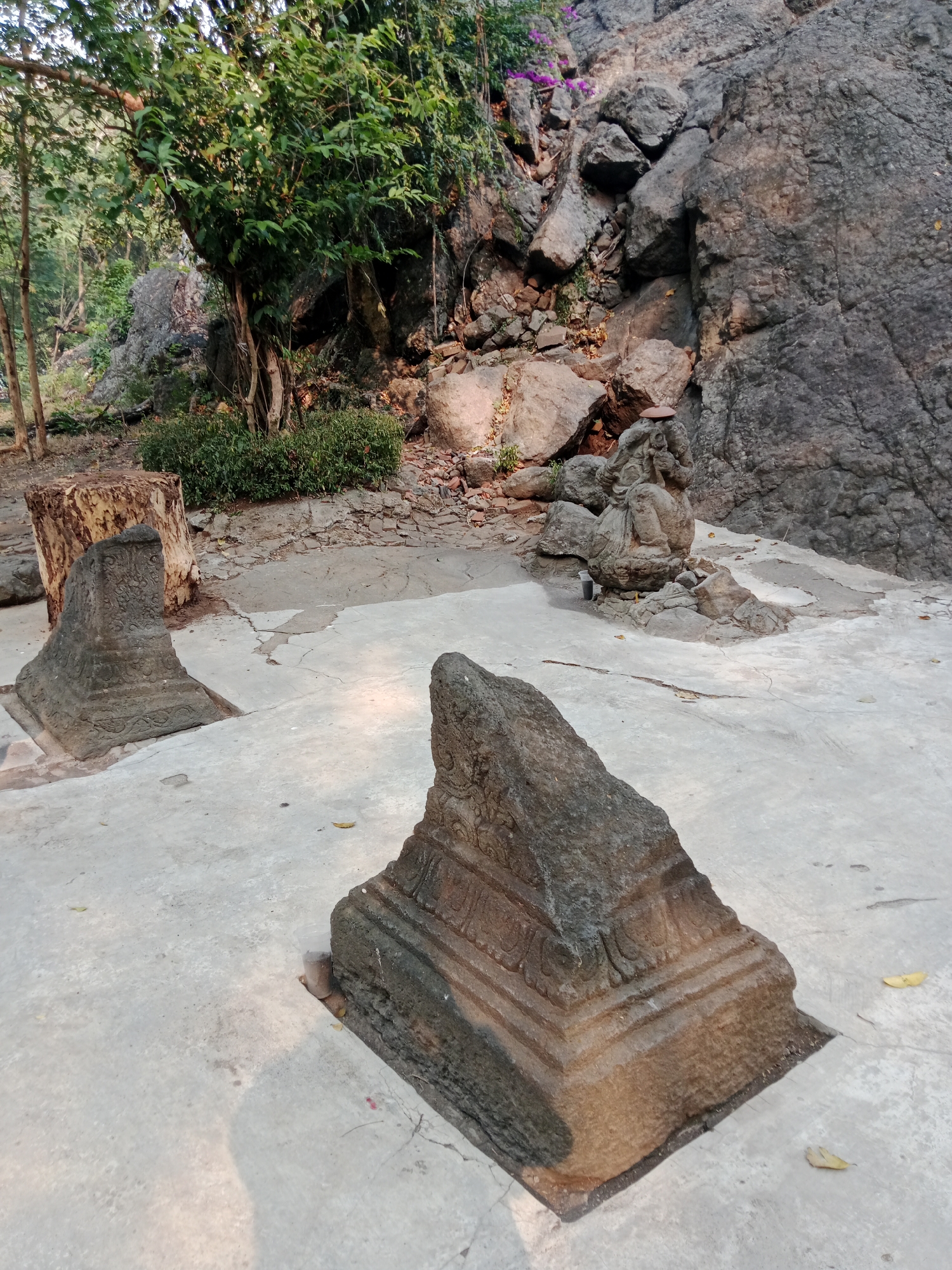 The image depicts an outdoor scene at a historical site, possibly a temple or ancient ruin, located in Kediri City, Indonesia. In the foreground, two weathered stone structures, likely architectural remnants, sit on a cracked concrete platform. The structure on the left is triangular in shape and exhibits intricate carvings, while the one on the right is partially visible and appears similar in style. Behind these, a larger stone monument carved in the likeness of a seated figure, possibly a deity or historical personage, is nestled amongst rocks and a clump of green bushes. The background reveals a steep, rocky cliff face, partially covered by foliage, with some purple flowers blooming near the top. Several trees, including a large one with dense green leaves, are also present, casting shadows on the ground. The overall environment suggests an ancient and somewhat overgrown historical park or archaeological site. There are no people visible in the scene.