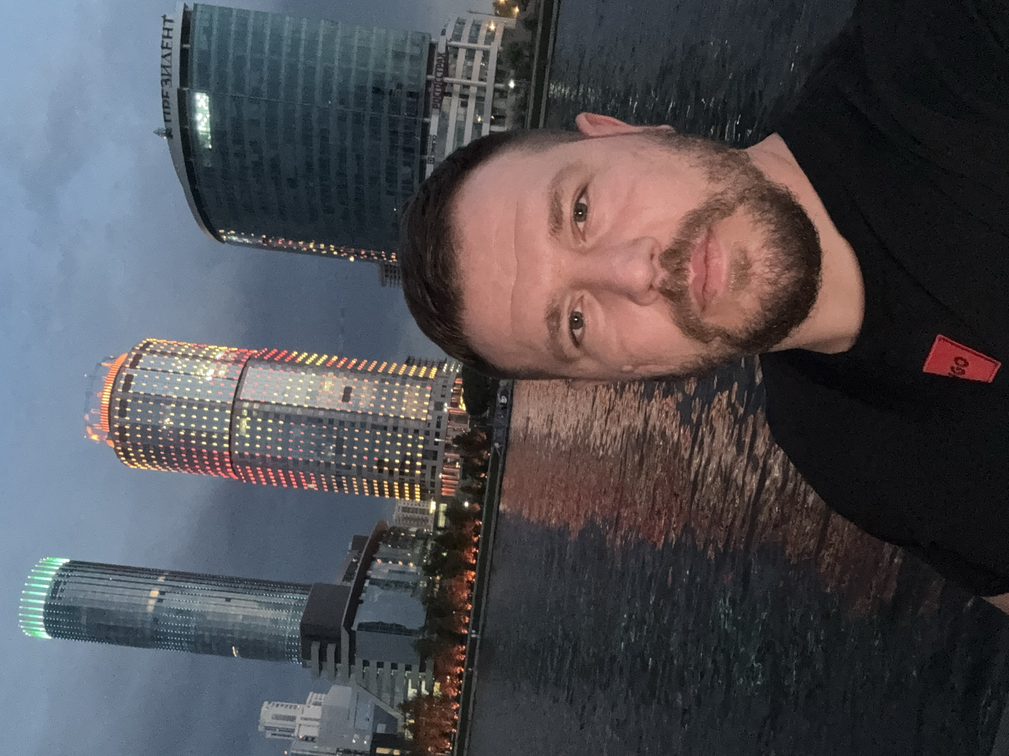 A man is taking a selfie against the backdrop of the cityscape of Yekaterinburg, Russia. The buildings visible in the background are prominent landmarks of the city. From top to bottom in the image, there is the "President" business center, the "Iset Tower," and the "Demidov Plaza" building. The man is wearing a black shirt with a red patch that seems to bear the letters "GO". The image captures a body of water reflecting the city lights, indicative of an evening scene.