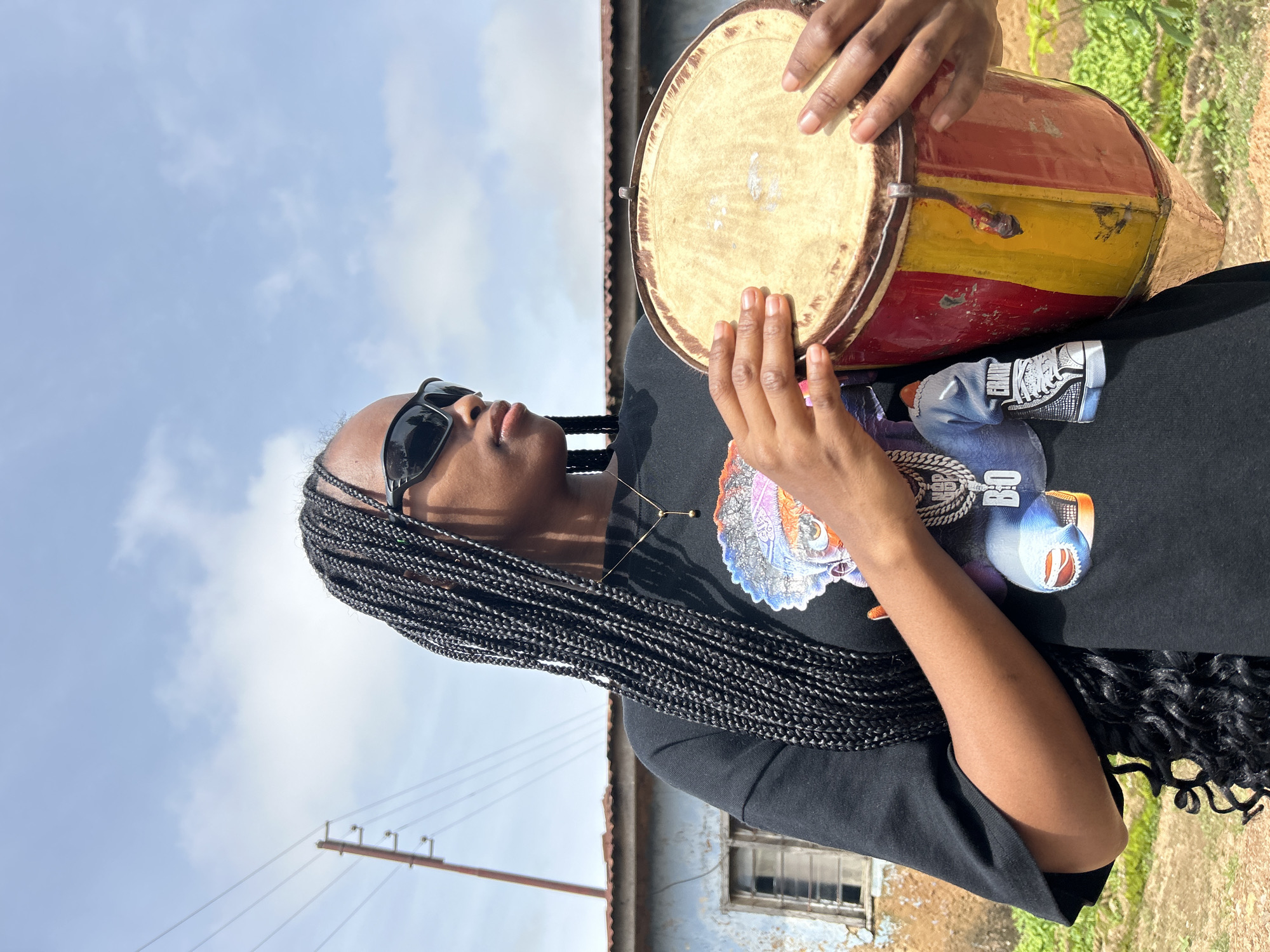 A person with braided hair and sunglasses is playing a drum outdoors. The drum is cylindrical with a light-colored head and a body painted in red and yellow stripes. The person's hands are positioned to strike the drum. They are wearing a black t-shirt with a graphic print featuring a colorful, abstract design that includes elements resembling animals and text. A thin chain with a pendant is visible around their neck.
The scene is set outdoors under a cloudy sky. In the background, there is a building with a corrugated metal roof and a window. A wooden pole with electrical wires is also visible in the distance. The overall impression is that of a casual, possibly warm day. The person's gaze is directed upwards, suggesting an engagement with the sky or the act of playing. The setting hints at a rural or semi-urban environment in Jos, Nigeria, given the building style and the presence of utility poles.