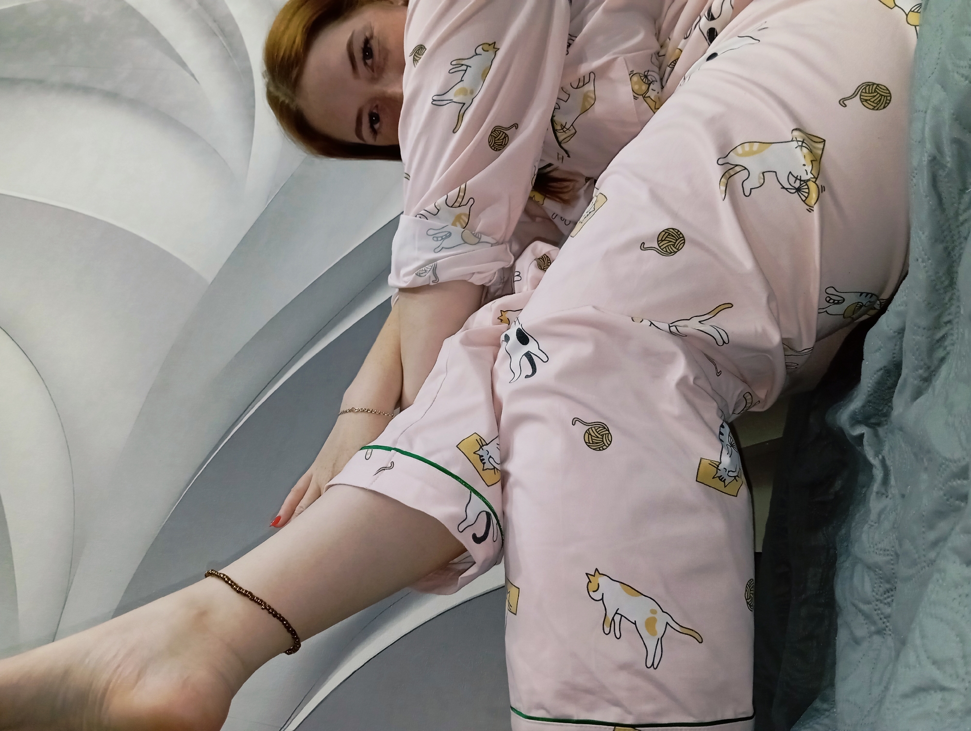 A woman is lying down, wearing pink pajamas with cat designs, on a bed or couch. The background includes a gray and white patterned wall or headboard and a blue patterned blanket. She is wearing an anklet and bracelet. The location is not apparent, and there is no indication it is Yurgamysh, Russia.