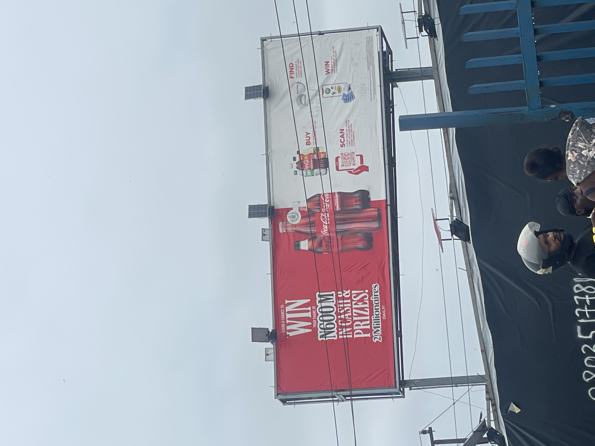 The image shows a Coca-Cola billboard in Lagos, Nigeria, advertising a promotion with N600 million in cash and prizes, featuring two millionaires daily. The billboard prompts people to buy, scan a code, and find information under the cap to win. A separate blue structure is visible next to the billboard, and there are people in the immediate vicinity, one wearing a white helmet. The sky is overcast.