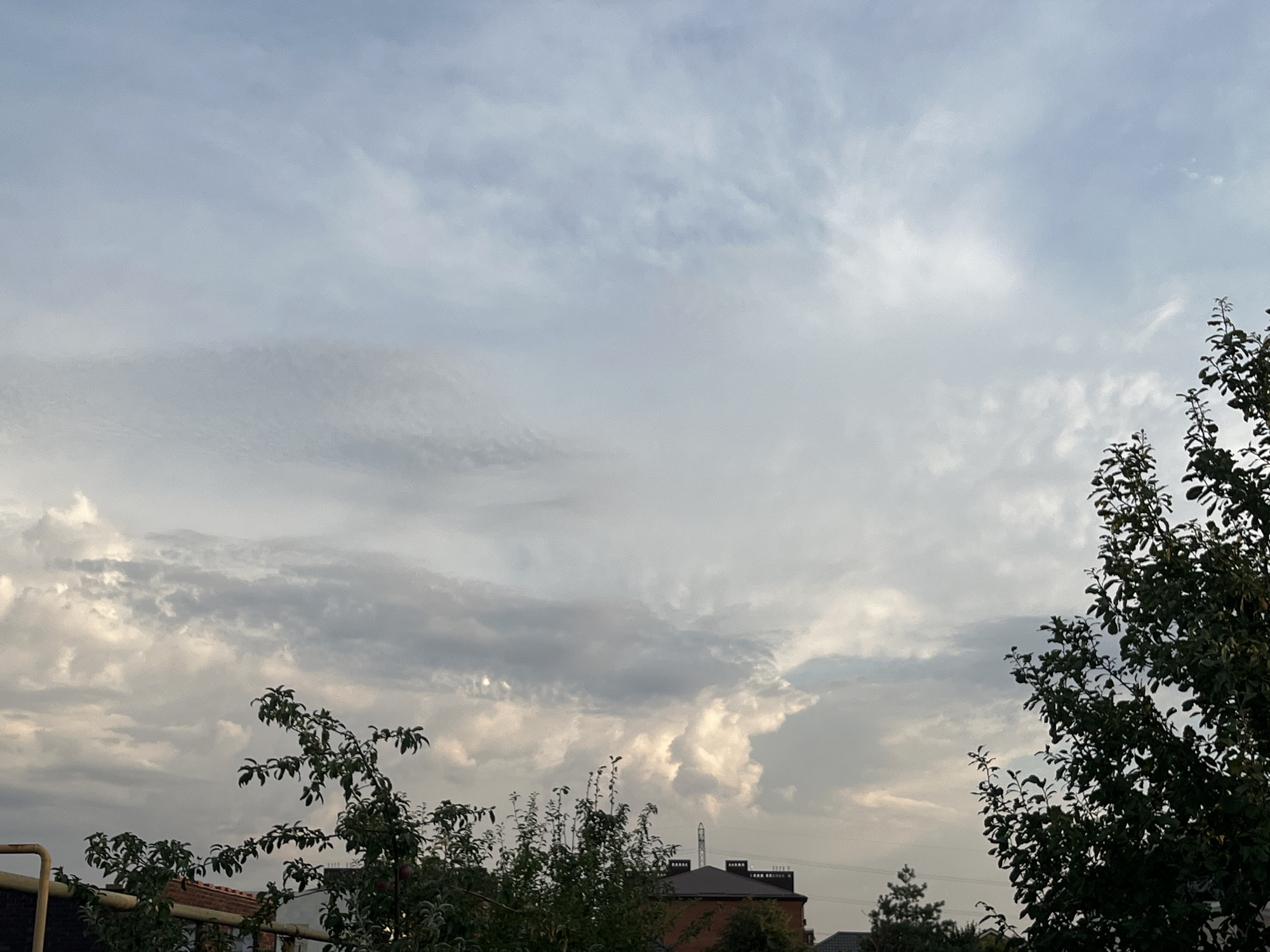 A broad, overcast sky dominates the frame, with pale blue and grey clouds forming a textured canopy. In the foreground, the tops of various trees and the roofs of buildings are visible, suggesting an urban or suburban setting. To the right, a large, dark green tree with dense foliage is silhouetted against the sky. On the left, a section of a yellow pipe extends diagonally upwards. The scene appears to be captured during daylight hours, possibly late afternoon or early evening, given the muted lighting.