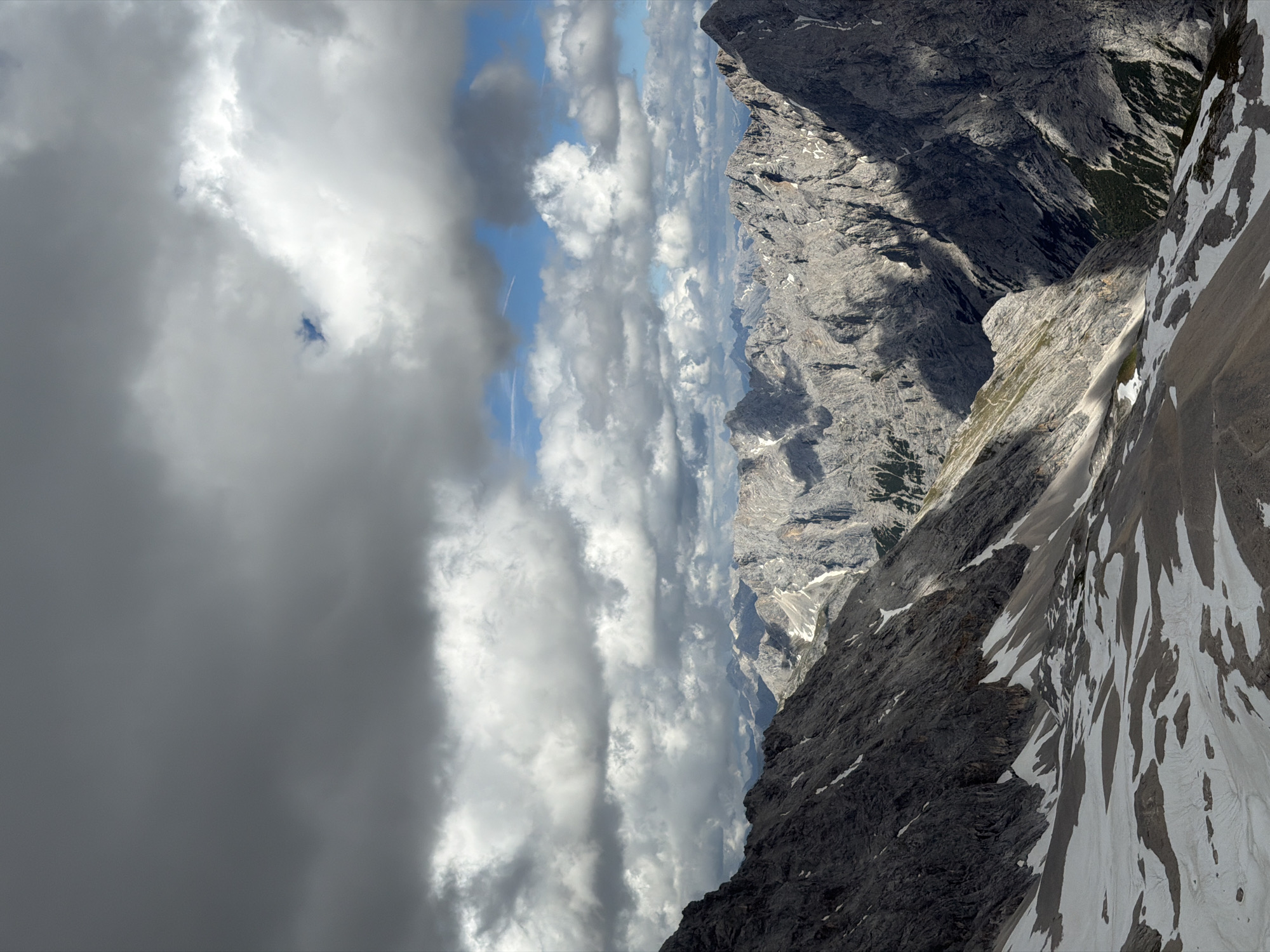 This media file captures a dramatic mountain landscape. The primary subjects are the rugged, grey, and textured mountain slopes, some of which are partially covered in snow. The scene is dominated by a vast sky filled with a mix of dark, heavy clouds and brighter, scattered white clouds. Gaps in the clouds reveal patches of blue sky, suggesting dynamic weather.
The perspective is from a high vantage point, looking down and across a steep mountain face. The time of day appears to be daytime, with sunlight intermittently breaking through the clouds, creating contrasting areas of light and shadow on the mountains. The weather is clearly variable, with a sense of impending or passing rain indicated by the dark clouds.
Notable details include the stark contrast between the dark, rocky foreground and the lighter, snow-dusted slopes further away. The intricate textures of the rock formations are prominent. The presence of snow suggests a high-altitude location, consistent with the context of Garmisch-Partenkirchen, Germany, a region known for its alpine scenery.
There is no visible text or people in the image. The overall mood conveyed is one of awe-inspiring natural beauty, with a touch of wildness and ruggedness due to the weather and terrain. The image evokes a sense of scale and the power of nature.